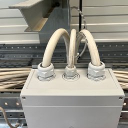 Goldfinger - Biała puszka elektryczna z trzema kablami w białej izolacji, zamontowana na metalowej konstrukcji w jasnym pomieszczeniu. Widoczne złącza kablowe.