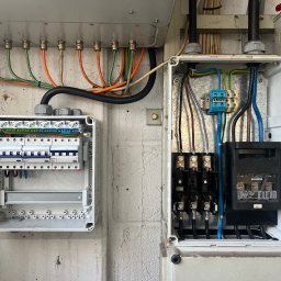 Goldfinger - Dwie otwarte skrzynki elektryczne z widocznymi przewodami, bezpiecznikami i elementami instalacji, zamontowane na ścianie. Widoczne kable w różnych kolorach.