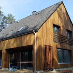 Nowoczesny dom jednorodzinny z elewacją z naturalnego drewna, widoczne rusztowania i materiały budowlane, dach z ciemnej dach&oacute;wki, okna z czarnymi ramami, częściowo zadaszony taras.