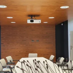 Sala konferencyjna z drewnianą boazerią na ścianie i suficie, białym stołem z mikrofonami oraz projektorem sufitowym.