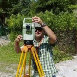 Mężczyzna w okularach przeciwsłonecznych ustawia teodolit na żółtym statywie na tle zieleni.