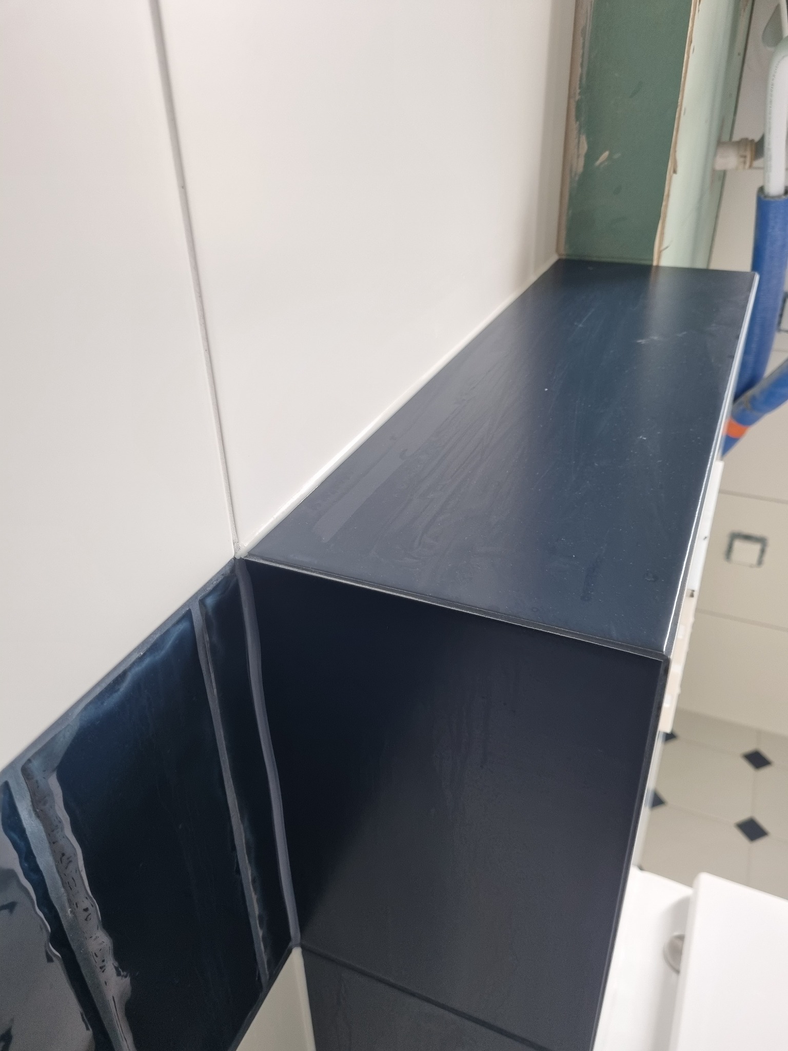 Półka z ciemnych płytek ceramicznych w łazience, kontrastująca z białymi płytkami ściennymi i widocznym fragmentem instalacji wodnej w tle.