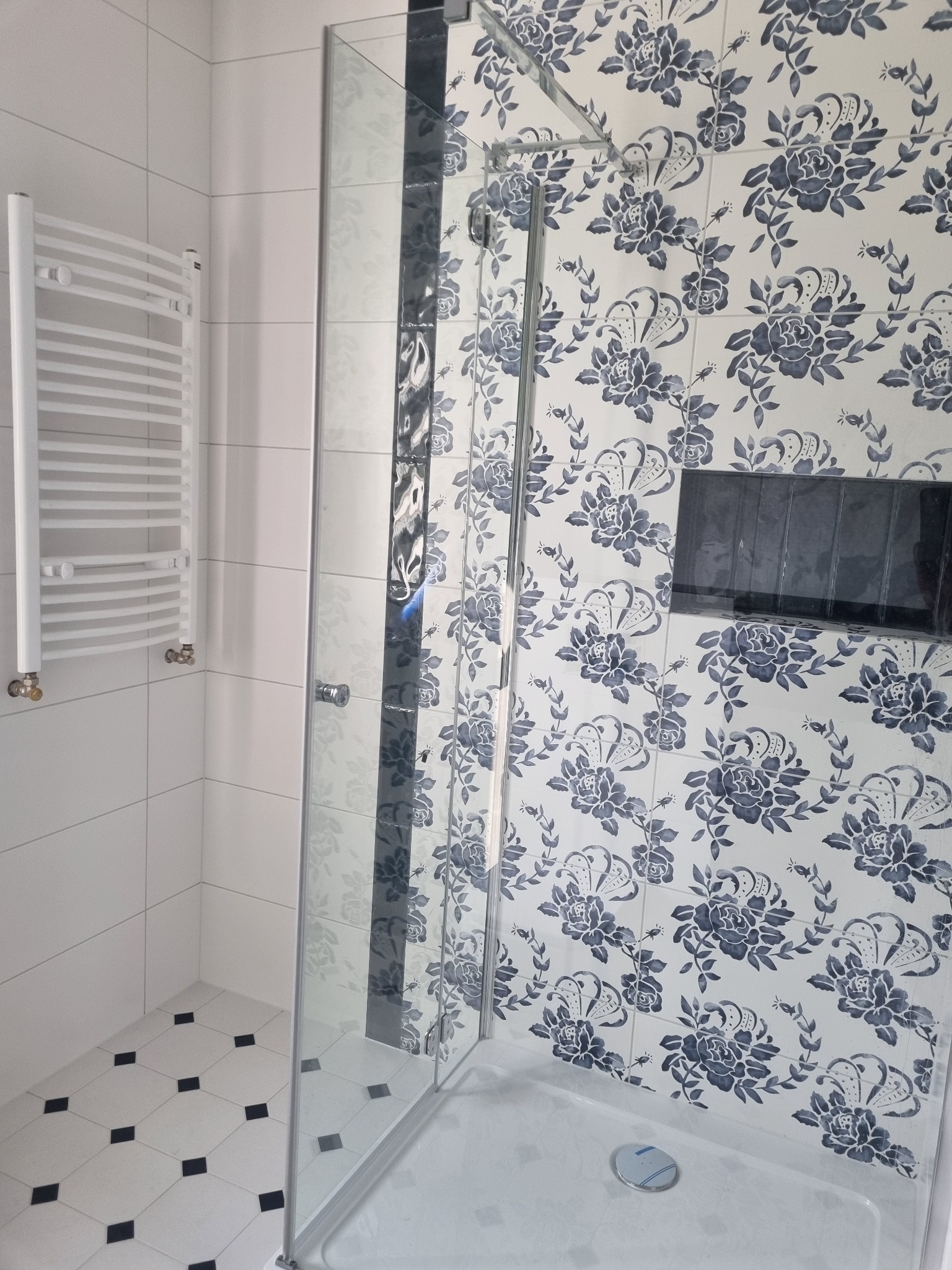 Kabina prysznicowa z białymi płytkami w kwiatowy wzór, grzejnik drabinkowy na ścianie i podłoga w biało-czarną szachownicę.