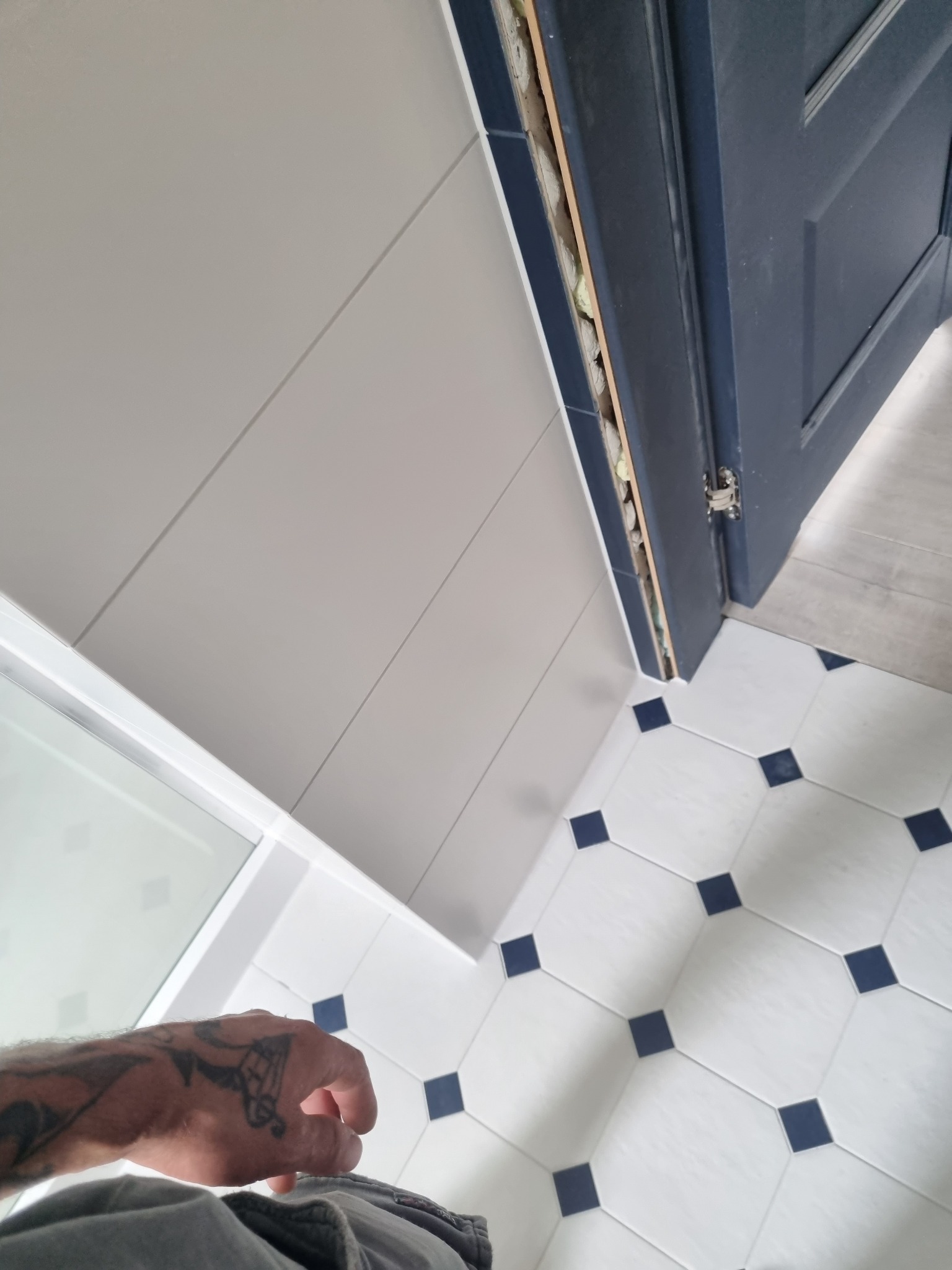 Biało-granatowa łazienka w trakcie remontu: widoczne panele ścienne, płytki podłogowe z granatowymi kwadratami, ościeżnica drzwi z pianką montażową i fragment ręki z tatuażem.