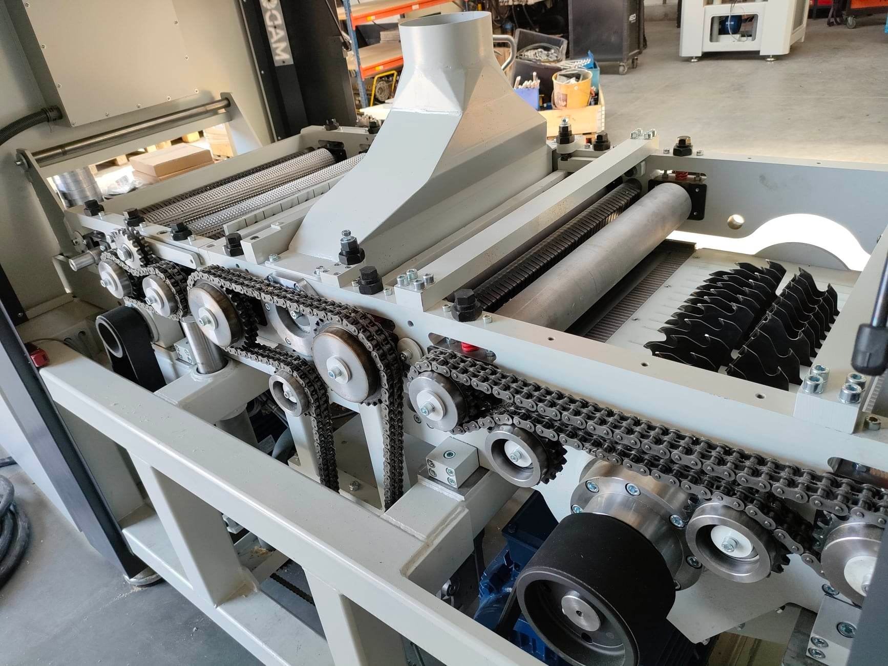 Strugarka dwustronna z wielopiłą MADORA DYRA 630, widoczne łańcuchy napędowe, wały i ostrza tnące, szara obudowa, oświetlenie warsztatowe.