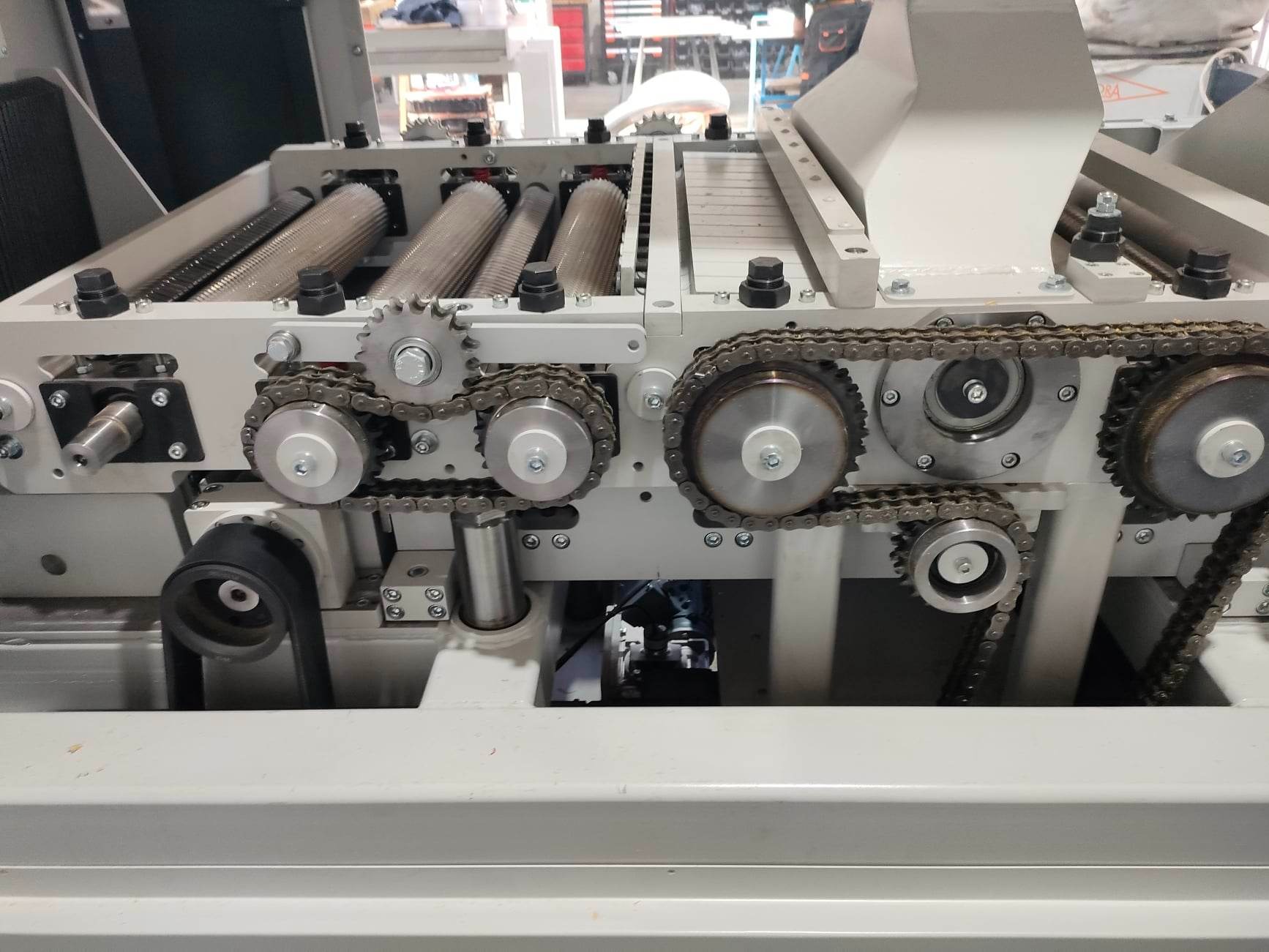 Strugarka dwustronna z wielopiłą, widoczne łańcuchy, zębatki i wałki podające materiał, detale mechanizmu posuwu.