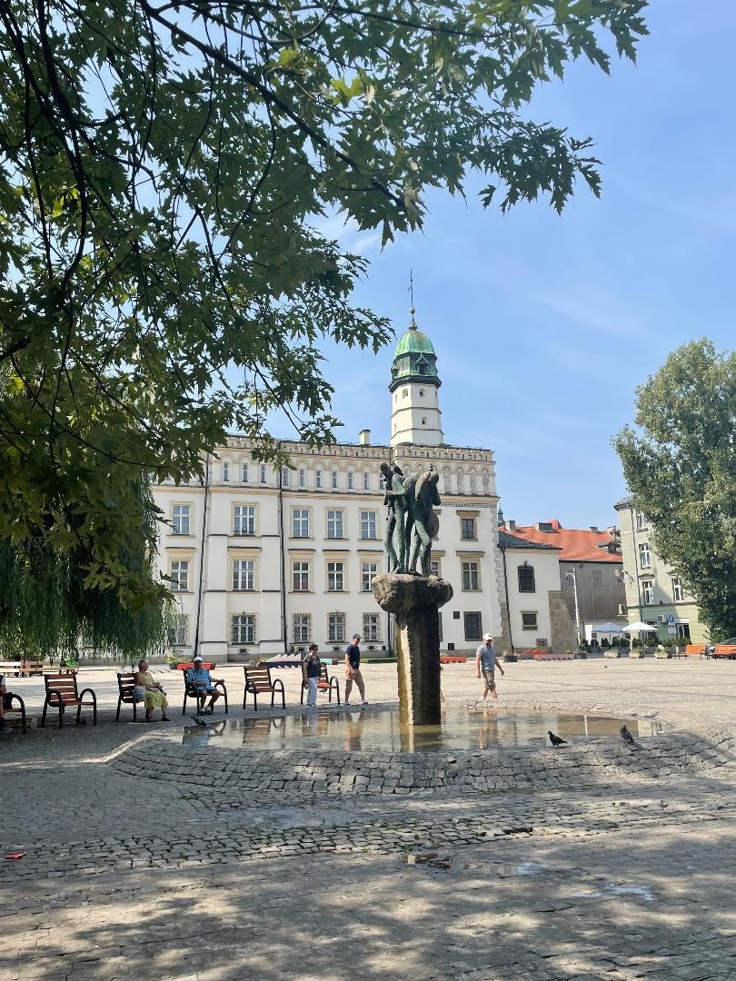 Rynek miejski z fontanną, budynkiem ratusza w tle i spacerującymi ludźmi w słoneczny dzień. Woda w fontannie odbija niebo. Na pierwszym planie kamienna nawierzchnia placu.