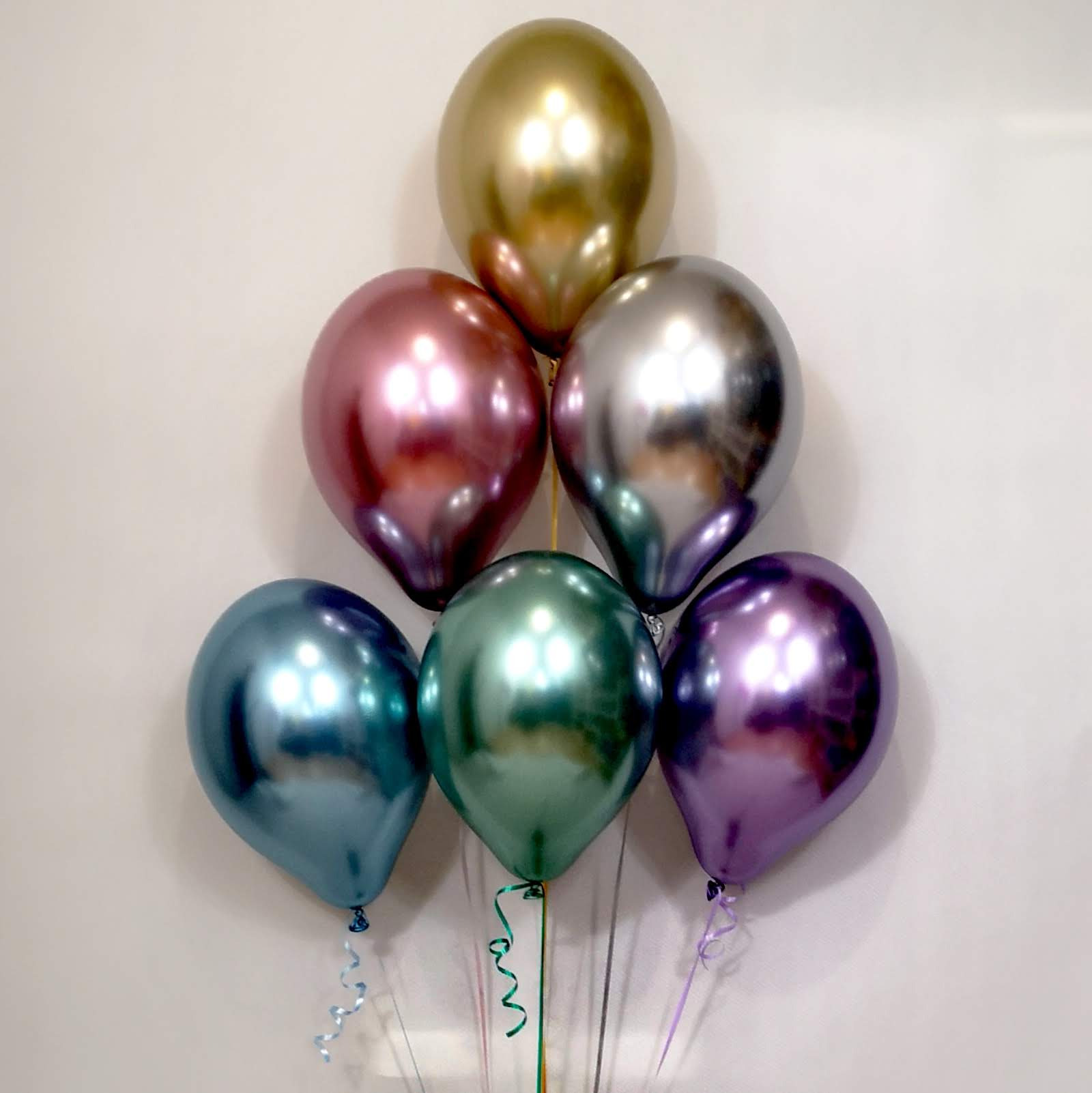 Bukiet siedmiu błyszczących, metalicznych balonów w kolorach złotym, różowym, srebrnym, niebieskim, zielonym i fioletowym, z kręconymi wstążkami, na tle jasnej ściany.