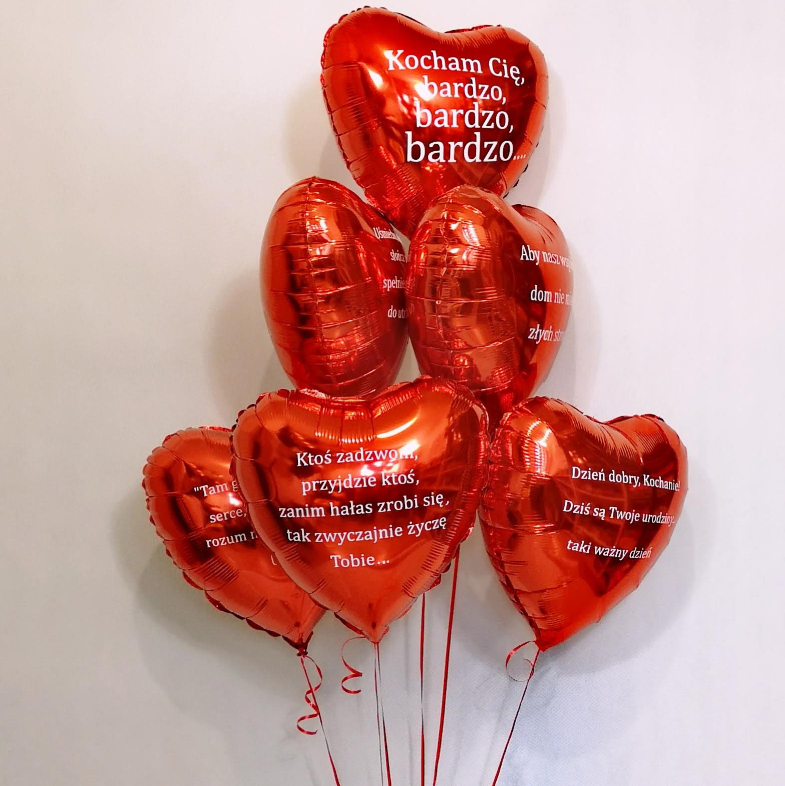 Bukiet czerwonych balonów w kształcie serc z białymi napisami z życzeniami urodzinowymi i miłosnymi, zawiązanych czerwonymi wstążkami na tle białej ściany.