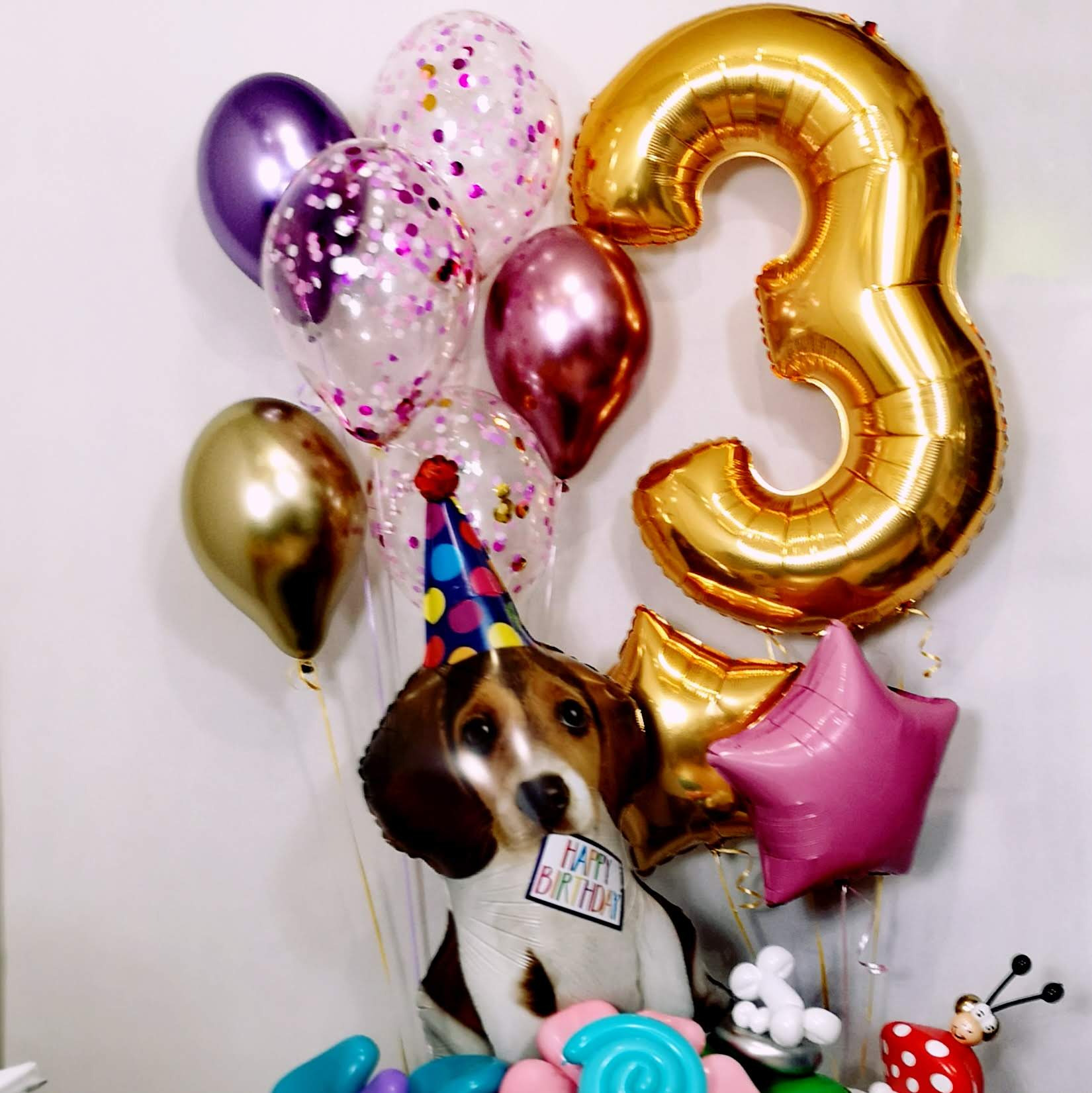 Dekoracja urodzinowa: balon w kształcie cyfry 3, balony z konfetti, balon foliowy w kształcie psa w czapeczce urodzinowej i z napisem 'Happy Birthday', różowe i złote gwiazdki.