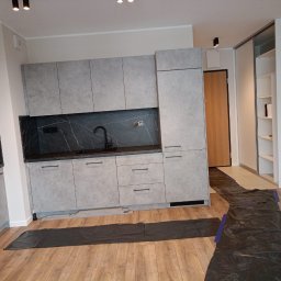 Profesjonalne Usługi Remontowo Wykończeniowe RUTEX - Nowoczesna kuchnia z szarymi matowymi szafkami i blatem z czarnego marmuru, czarna bateria, podłoga wyłożona panelami, zabezpieczona folią ochronną podczas prac.