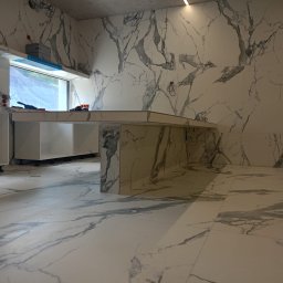 Profesjonalne Usługi Remontowo Wykończeniowe RUTEX - Nowoczesna kuchnia w trakcie wykańczania, z białymi szafkami bez frontów i blatem w marmurowy wzór, płytki podłogowe i ścienne również w marmurze, widoczne niedokończone instalacje elektryczne.