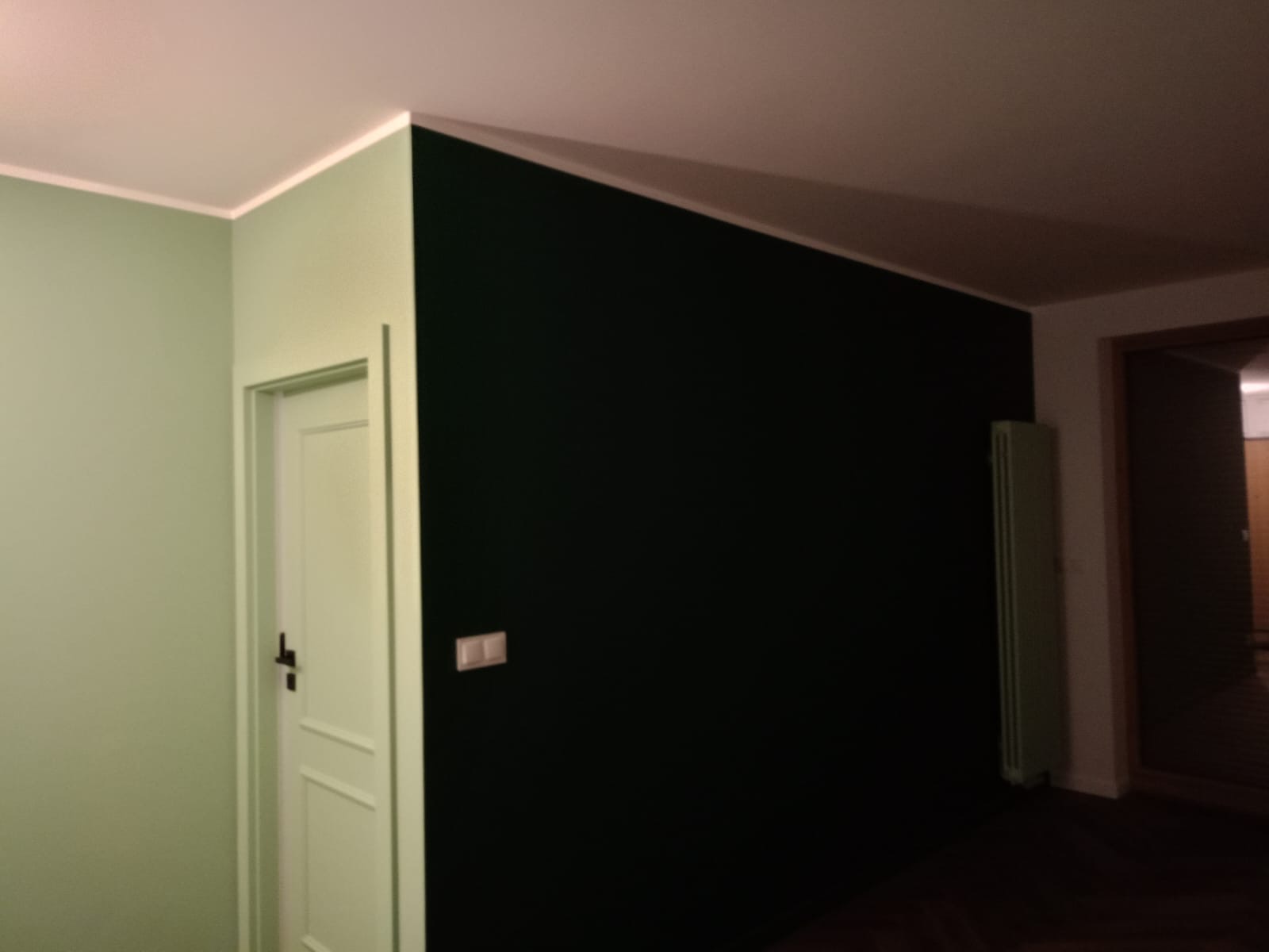 Ściana pomalowana na ciemnozielony kolor w zestawieniu z pistacjową ścianą i białą stolarką drzwiową, widoczny grzejnik w tle.
