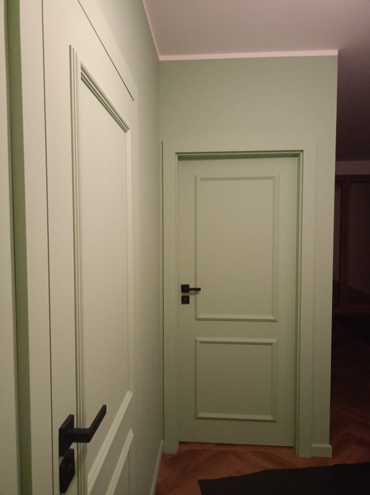 Dwa wewnętrzne, pomalowane na jasnozielono drzwi z czarnymi klamkami w korytarzu z parkietem ułożonym w jodełkę.
