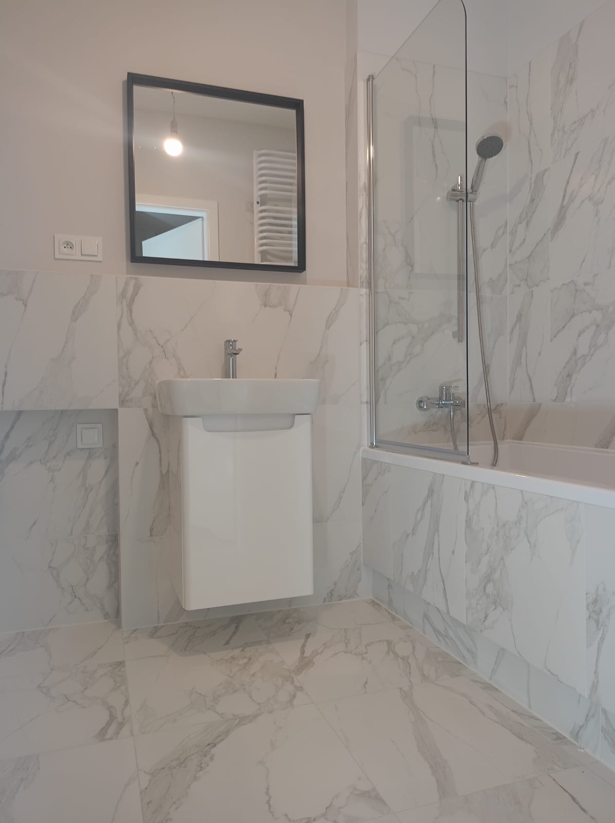 Nowoczesna łazienka z marmurowymi płytkami, białą szafką pod umywalką i kabiną prysznicową ze szklanym panelem.