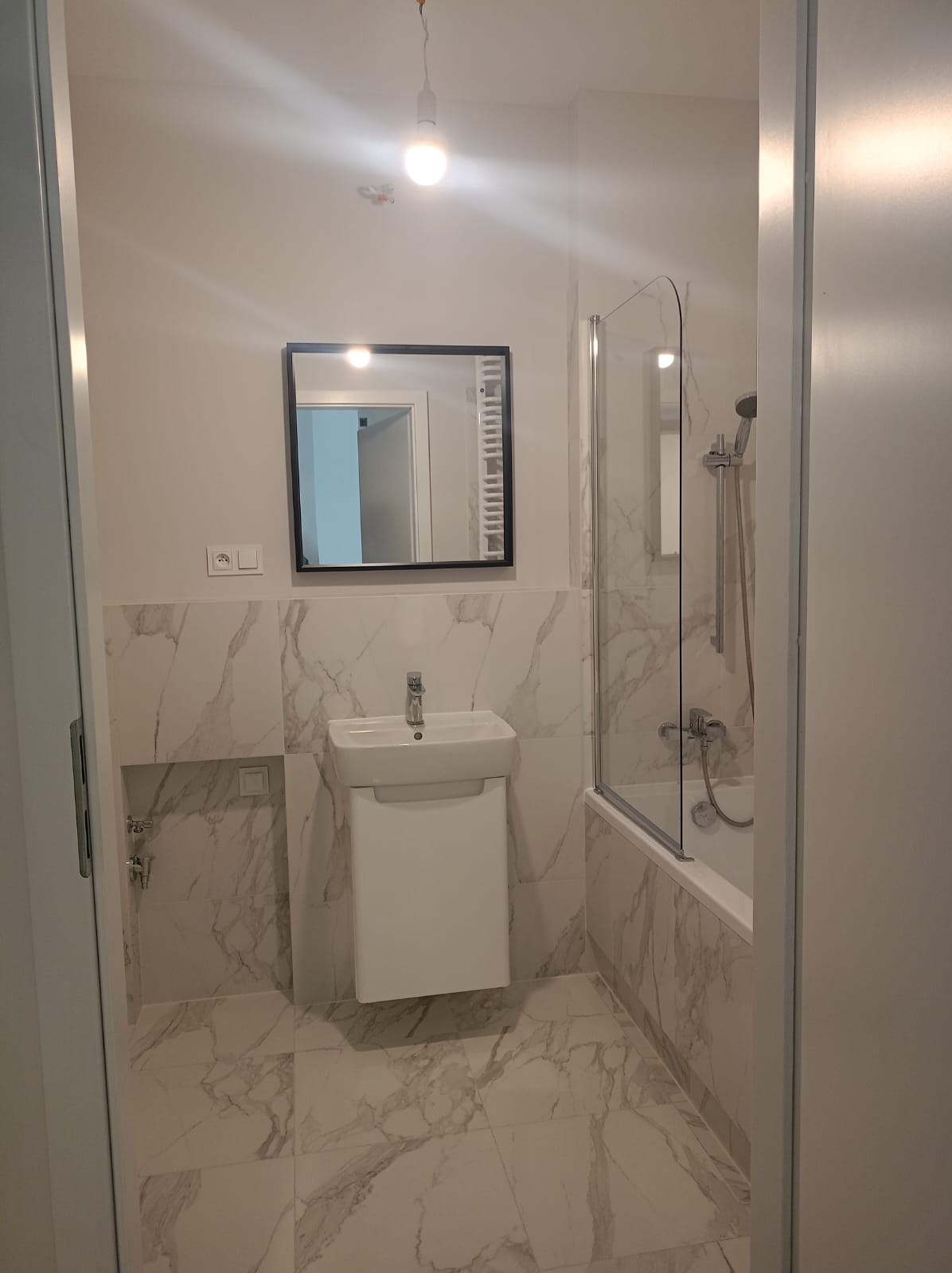 Nowoczesna łazienka z marmurowymi płytkami, wiszącą białą szafką z umywalką, lustrem w czarnej ramie i przeszklonym parawanem nawannowym. Widoczny surowy żyrandol z żarówką bez klosza.