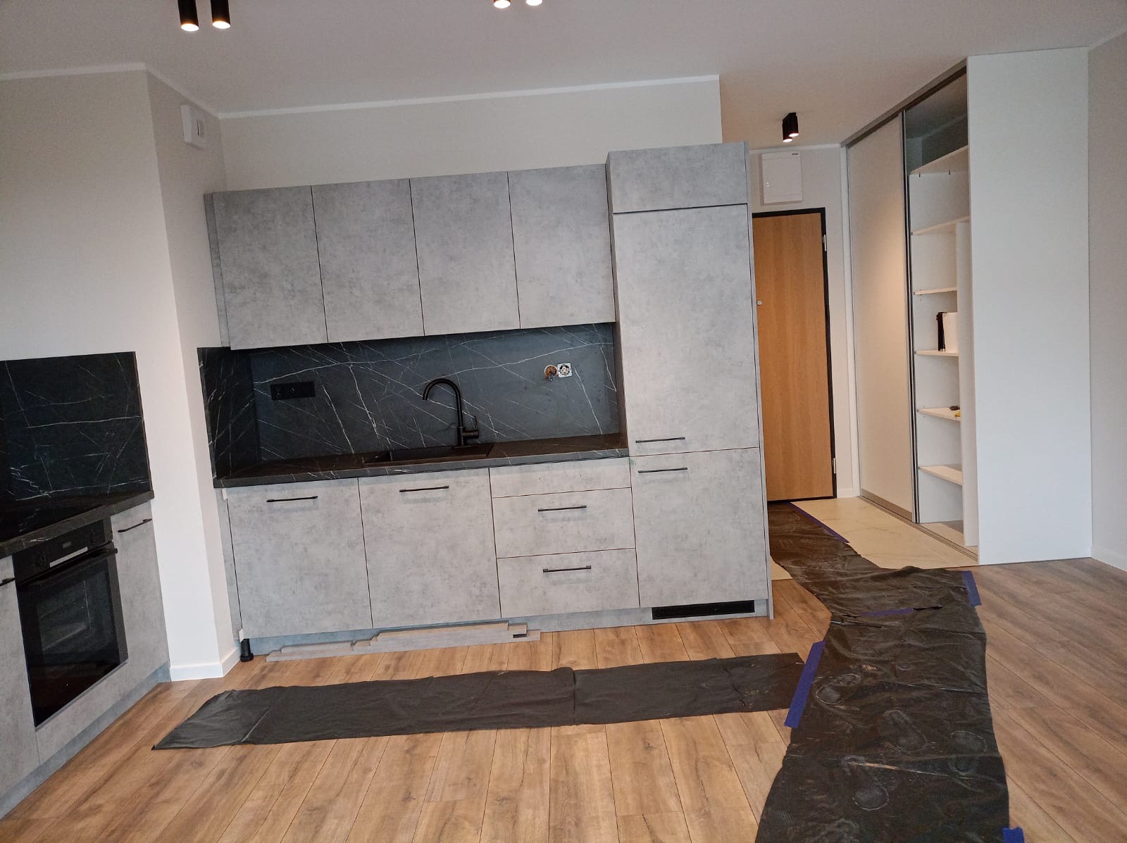 Nowoczesna kuchnia z szarymi matowymi szafkami i blatem z czarnego marmuru, czarna bateria, podłoga wyłożona panelami, zabezpieczona folią ochronną podczas prac.