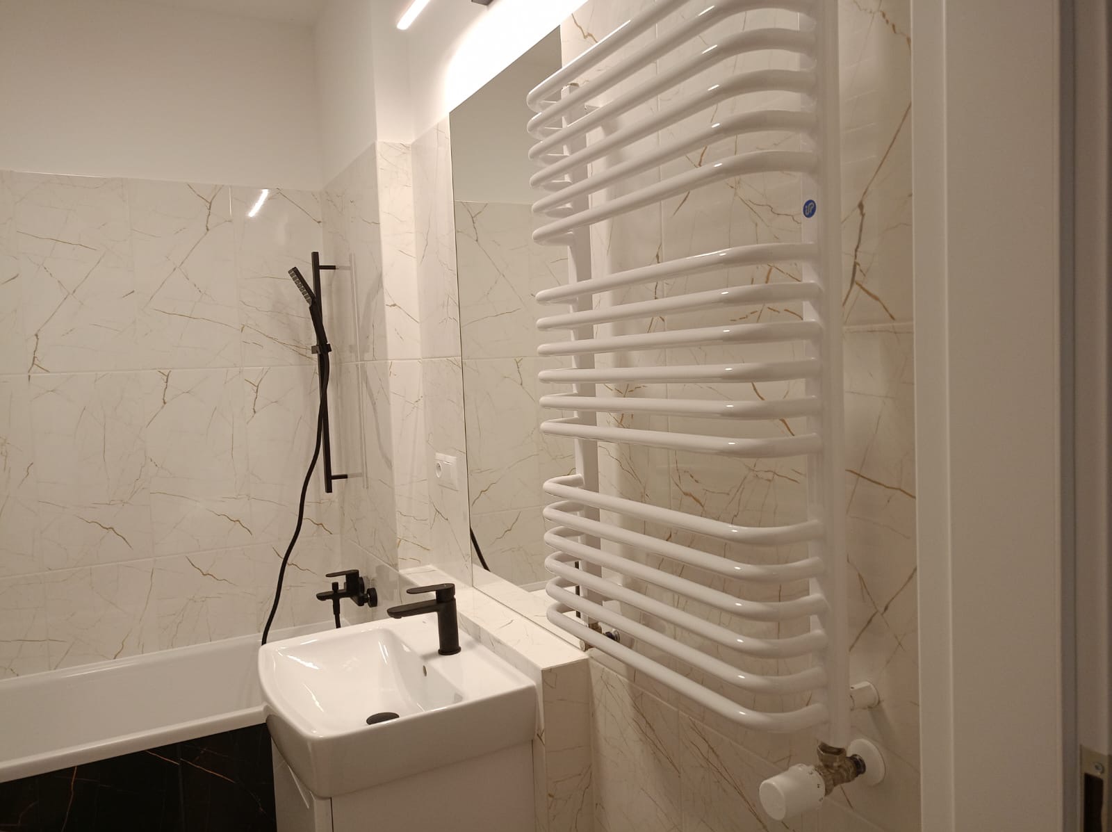 Nowoczesna łazienka z umywalką nablatową, czarną armaturą i białym grzejnikiem drabinkowym na tle płytek imitujących marmur.