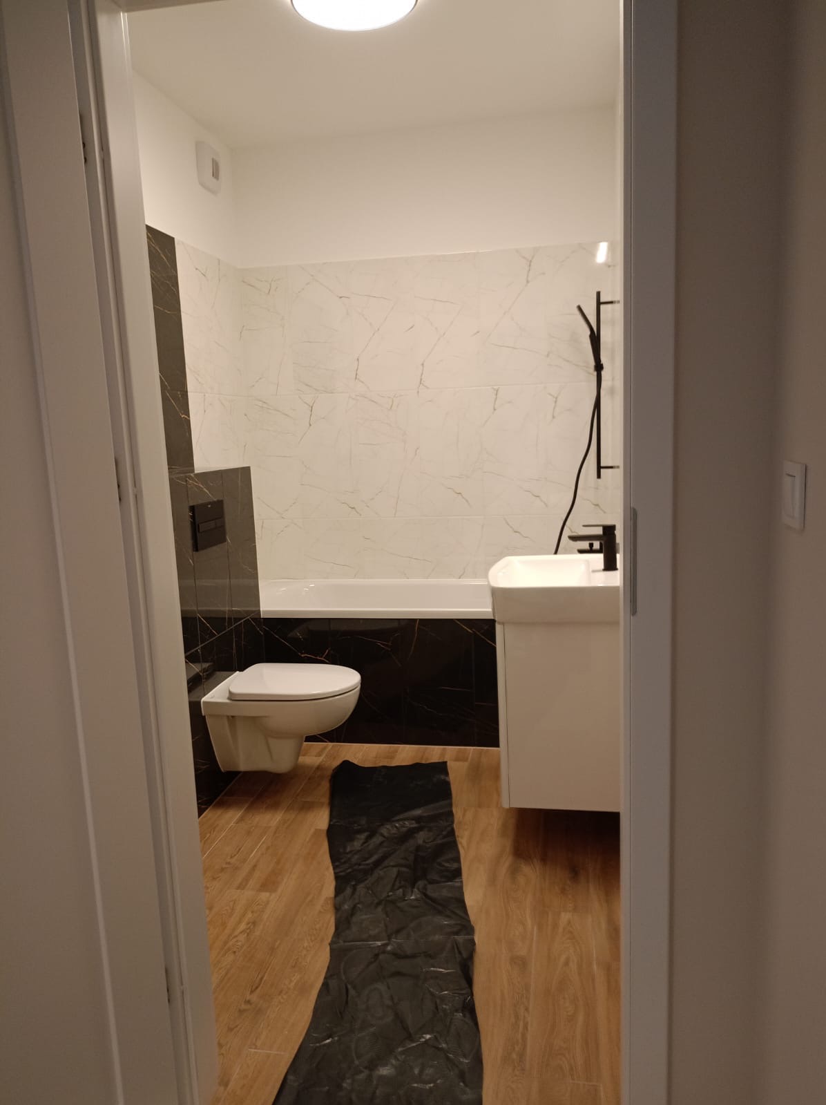 Nowoczesna łazienka z marmurowymi płytkami, czarną armaturą i drewnopodobną podłogą, zabezpieczona czarną folią ochronną.