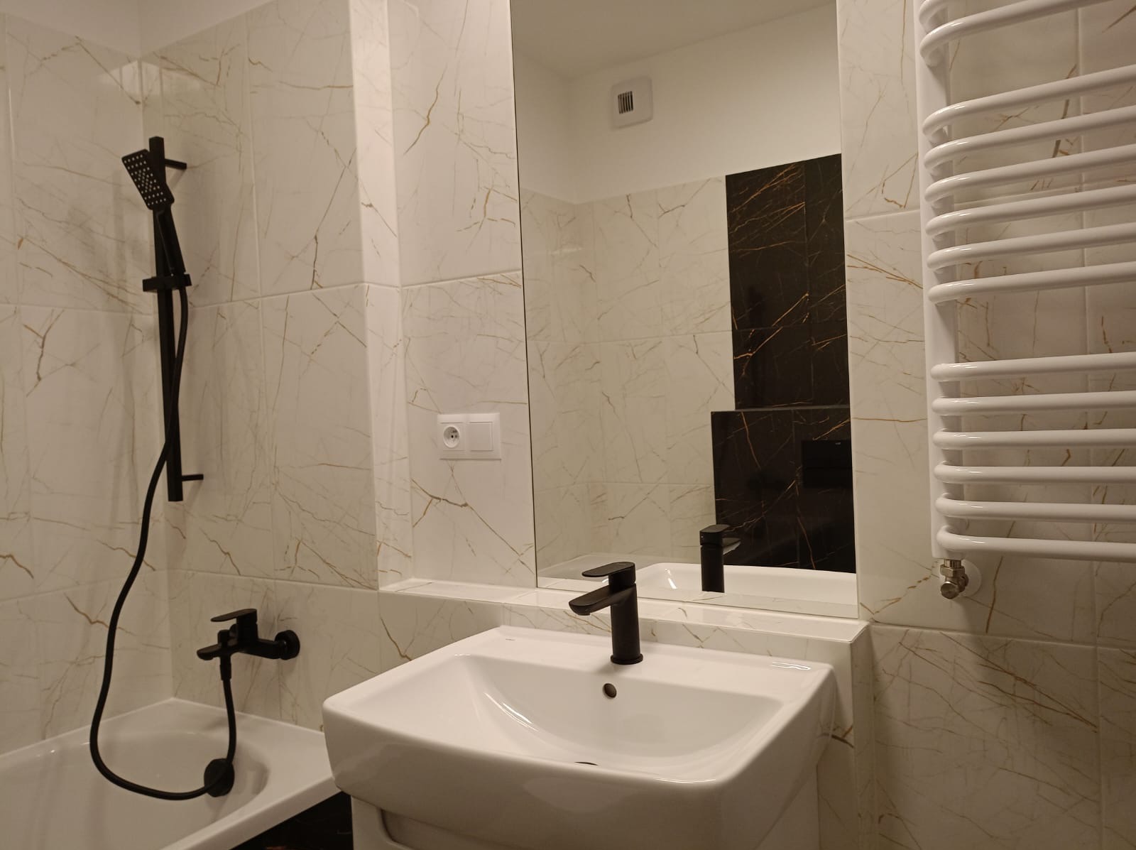 Wykończona łazienka z białymi marmurkowymi płytkami, czarną armaturą, białą umywalką i grzejnikiem drabinkowym.