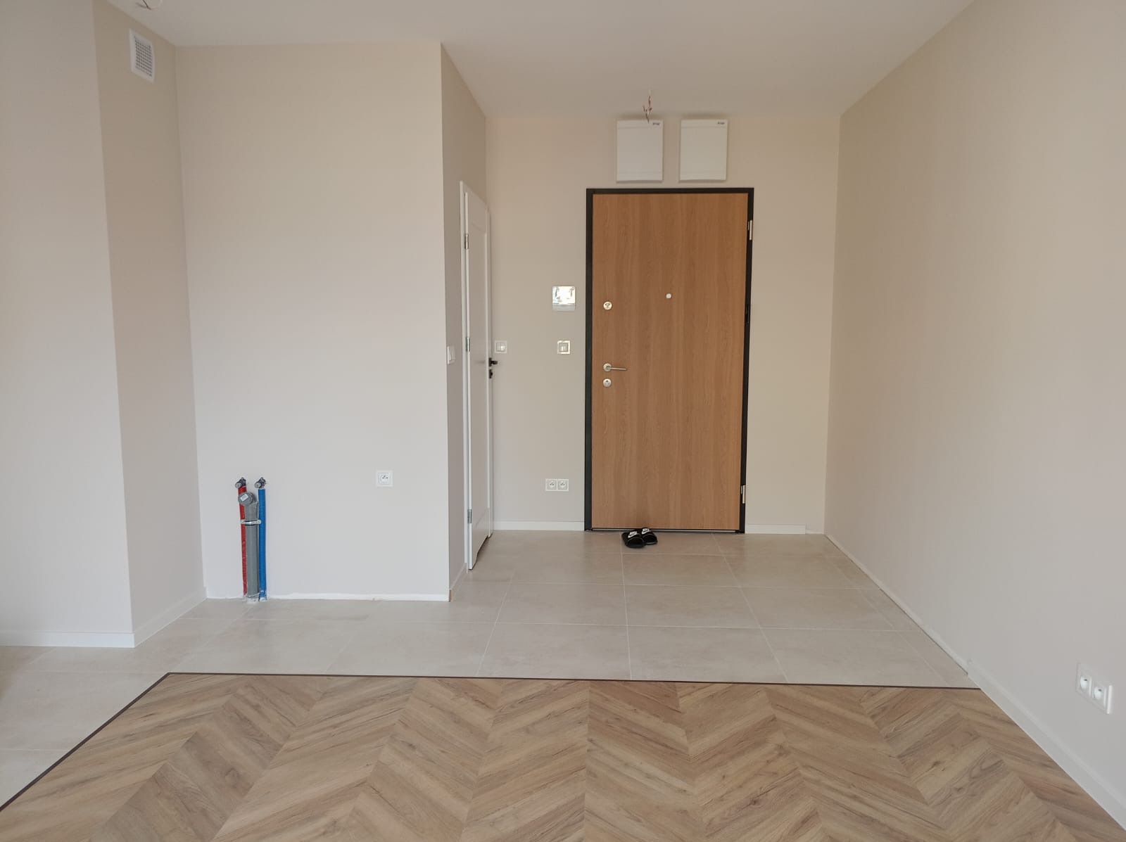 Nowe mieszkanie po remoncie: parkiet ułożony w jodełkę francuską przechodzi w jasne, kwadratowe płytki, widoczne drzwi wejściowe z czarną ramą i wewnętrzne, ściany pomalowane na beżowo, brak mebli.