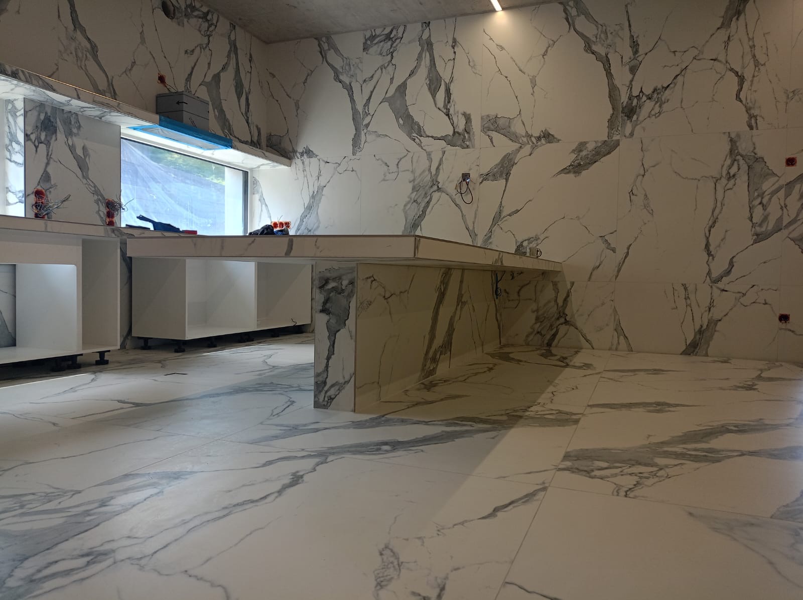 Nowoczesna kuchnia w trakcie wykańczania, z białymi szafkami bez frontów i blatem w marmurowy wzór, płytki podłogowe i ścienne również w marmurze, widoczne niedokończone instalacje elektryczne.