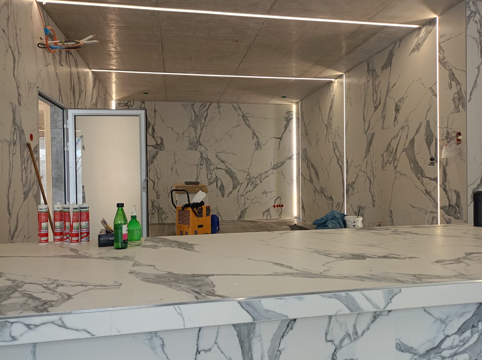 Wnętrze w trakcie wykańczania: marmurowe płytki na ścianach, oświetlenie LED w suficie i pionowe pasy świetlne, widoczne materiały budowlane i narzędzia na blacie.
