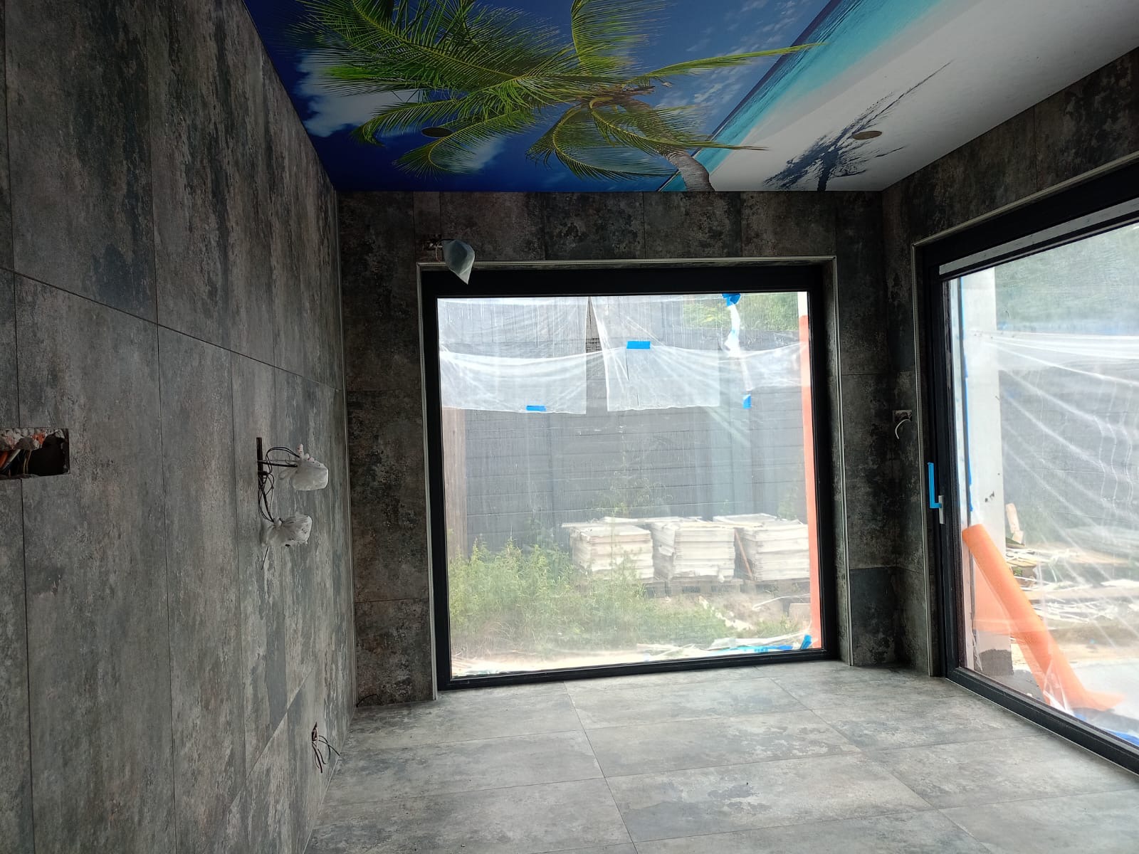 Łazienka w trakcie remontu: szare płytki imitujące kamień na ścianach i podłodze, sufit z fototapetą przedstawiającą plażę z palmami, okna zabezpieczone folią malarską, widoczne puszki elektryczne...