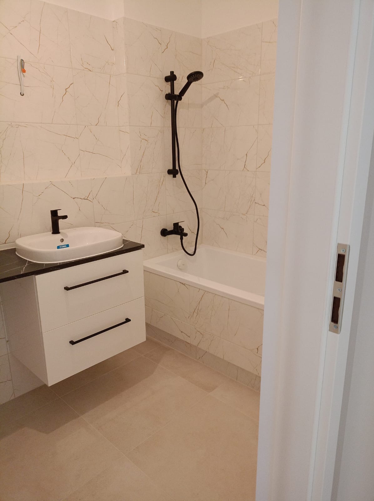 Nowoczesna łazienka z białą armaturą i czarnymi akcentami: bateria umywalkowa, deszczownica i uchwyty szafki pod umywalką. Płytki ścienne imitujące marmur.