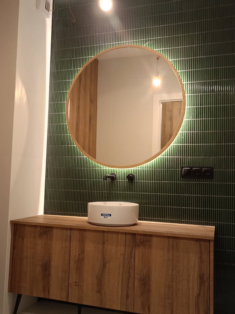 Nowoczesna łazienka z zieloną, pionowo ułożoną glazurą, okrągłym lustrem z podświetleniem LED, białą umywalką nablatową i szafką z drewnianym fornirem.