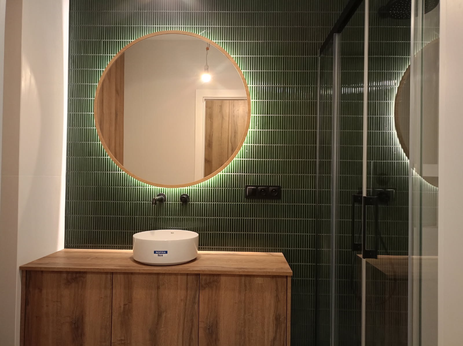 Nowoczesna łazienka z zielonymi, pionowymi płytkami, okrągłym lustrem z podświetleniem LED, drewnianą szafką pod umywalką i czarną armaturą.