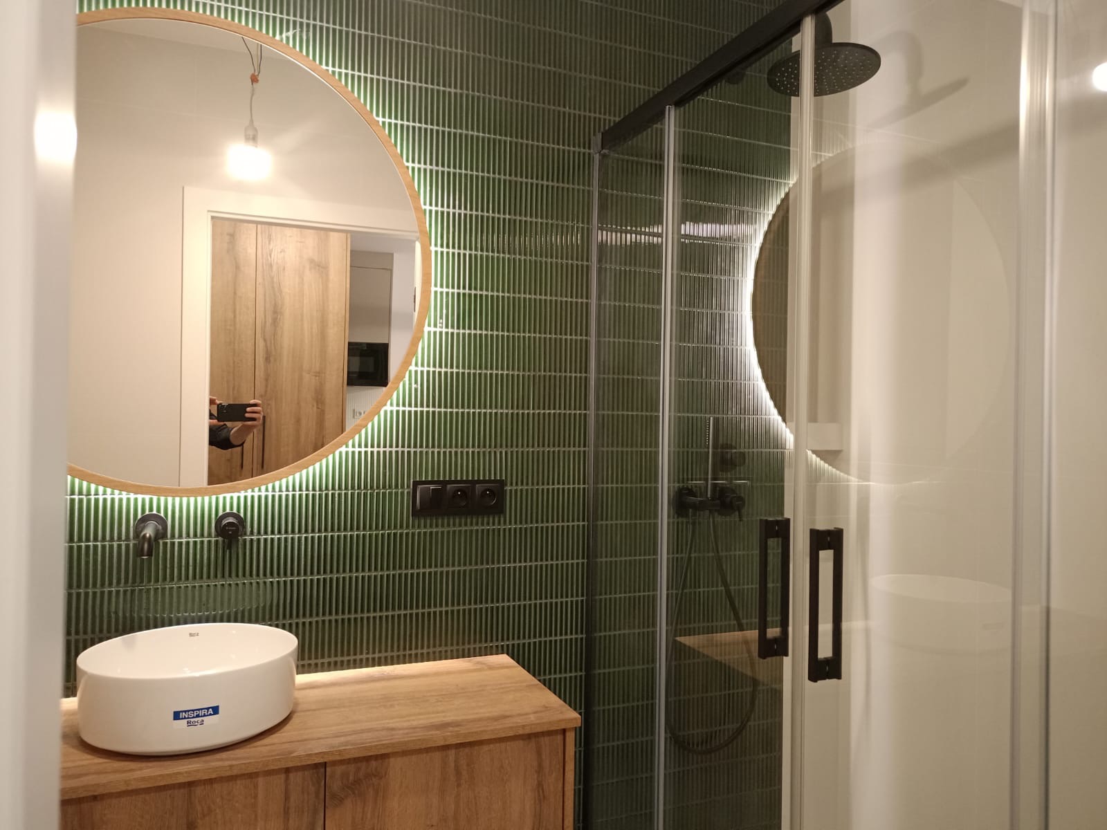 Nowoczesna łazienka z zielonymi, pionowo ułożonymi płytkami, okrągłe lustro z podświetleniem LED, drewniana szafka pod umywalką i kabina prysznicowa z czarnymi elementami.