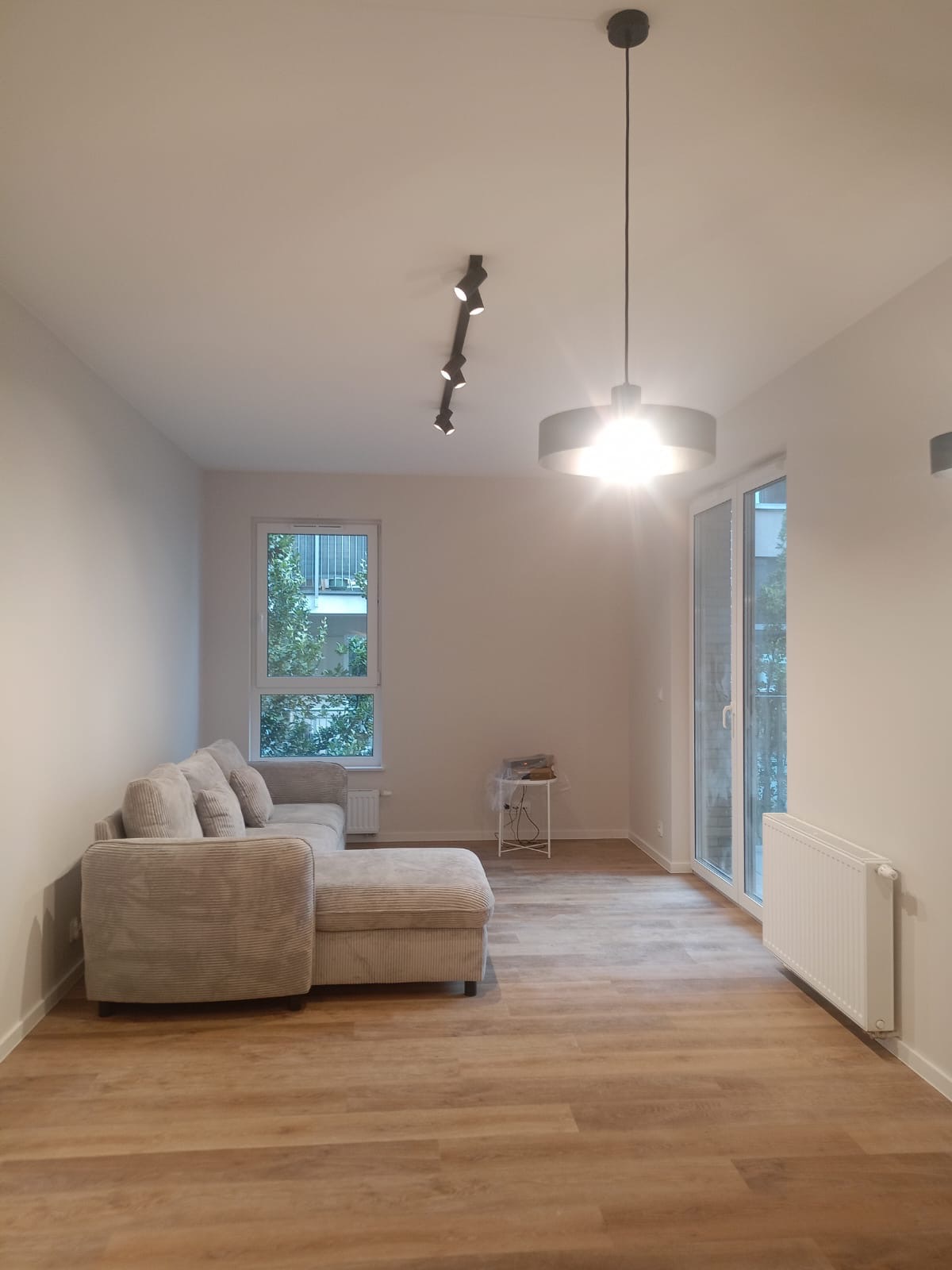 Jasne wnętrze salonu z drewnianą podłogą, beżową sofą narożną i nowoczesnym oświetleniem sufitowym. Widoczne okno i drzwi balkonowe.