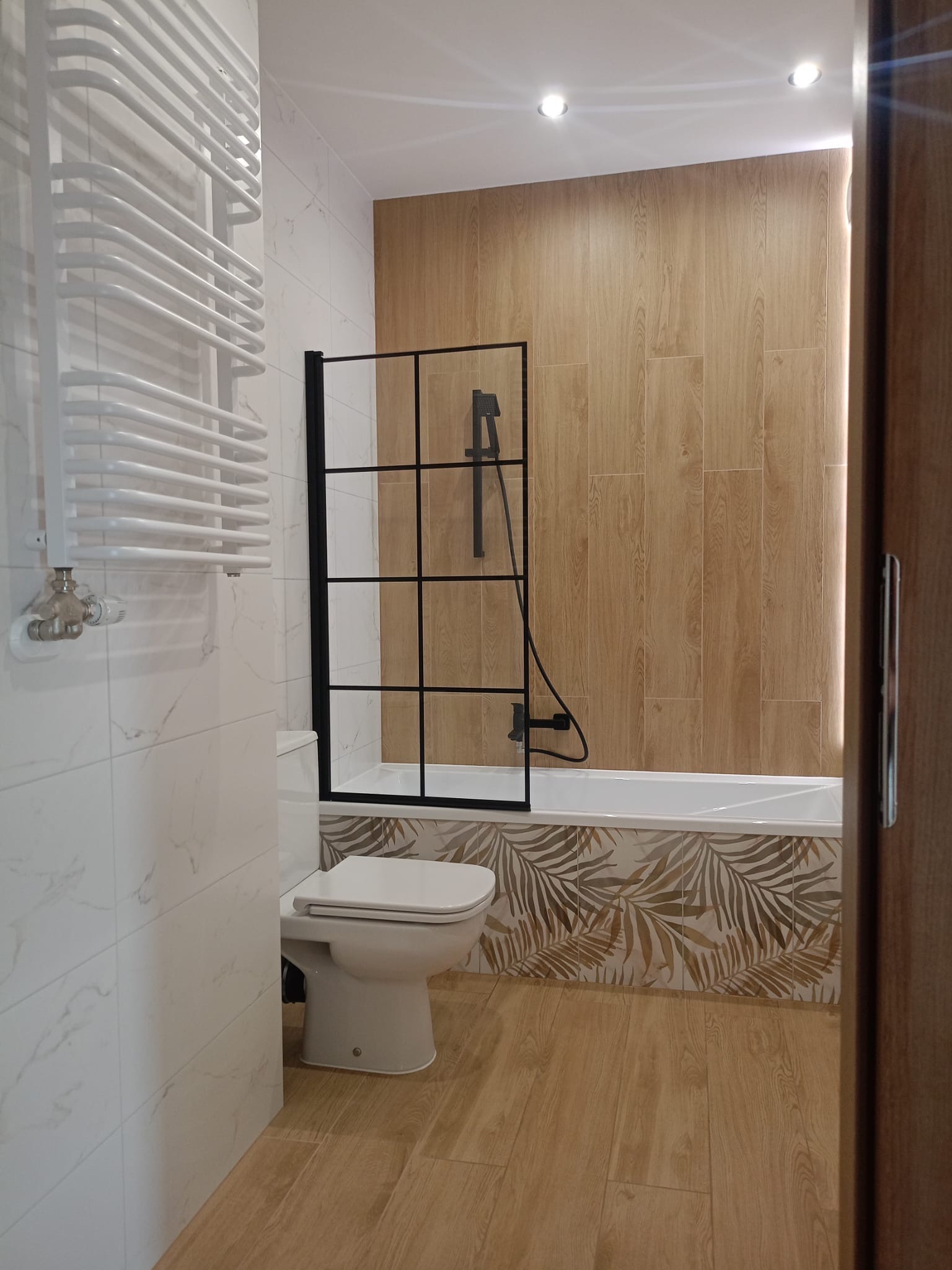 Nowoczesna łazienka z kabiną prysznicową typu walk-in, drewnopodobnymi płytkami i grzejnikiem drabinkowym. Uwagę zwracają płytki z motywem liści palm.