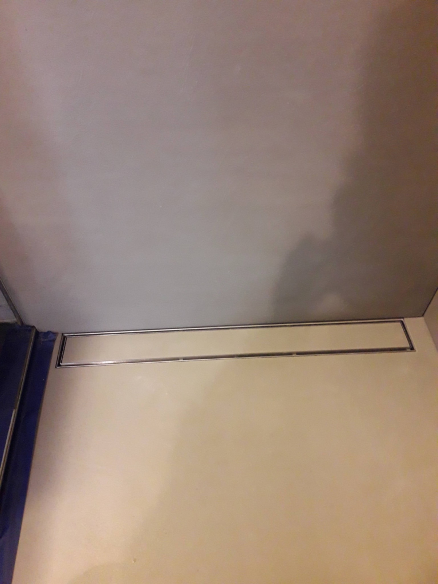 Zamontowany liniowy odpływ prysznicowy w jasnej, wykafelkowanej łazience, widoczne fragmenty niebieskiej obudowy.