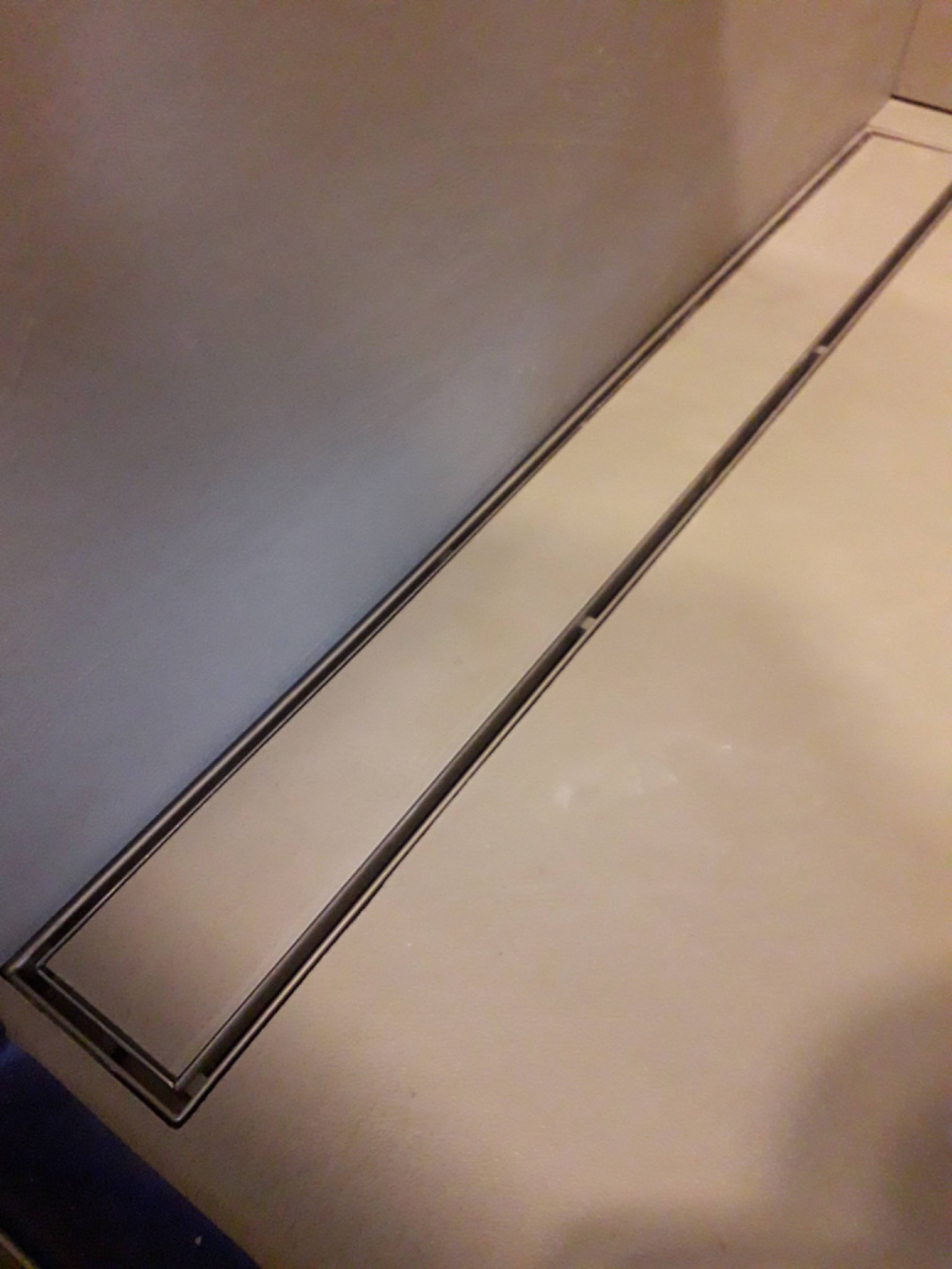 Dyskretny odpływ liniowy ze stali nierdzewnej w posadzce łazienki, przylegający do szarej ściany.