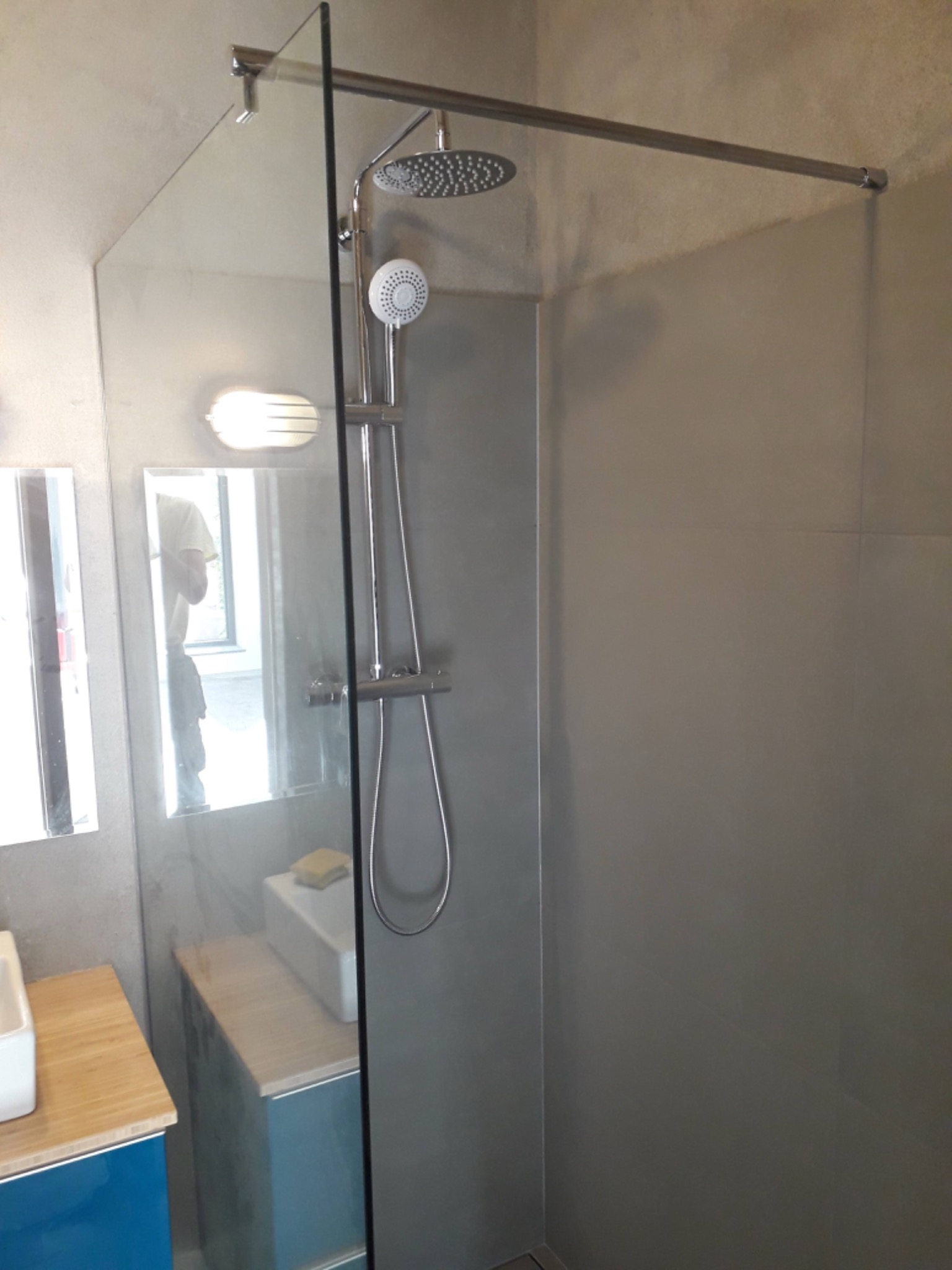 Nowoczesna kabina prysznicowa typu walk-in z deszczownicą i słuchawką, szara ściana z płytek wielkoformatowych, widoczne fragmenty szafek łazienkowych z blatem z jasnego drewna.