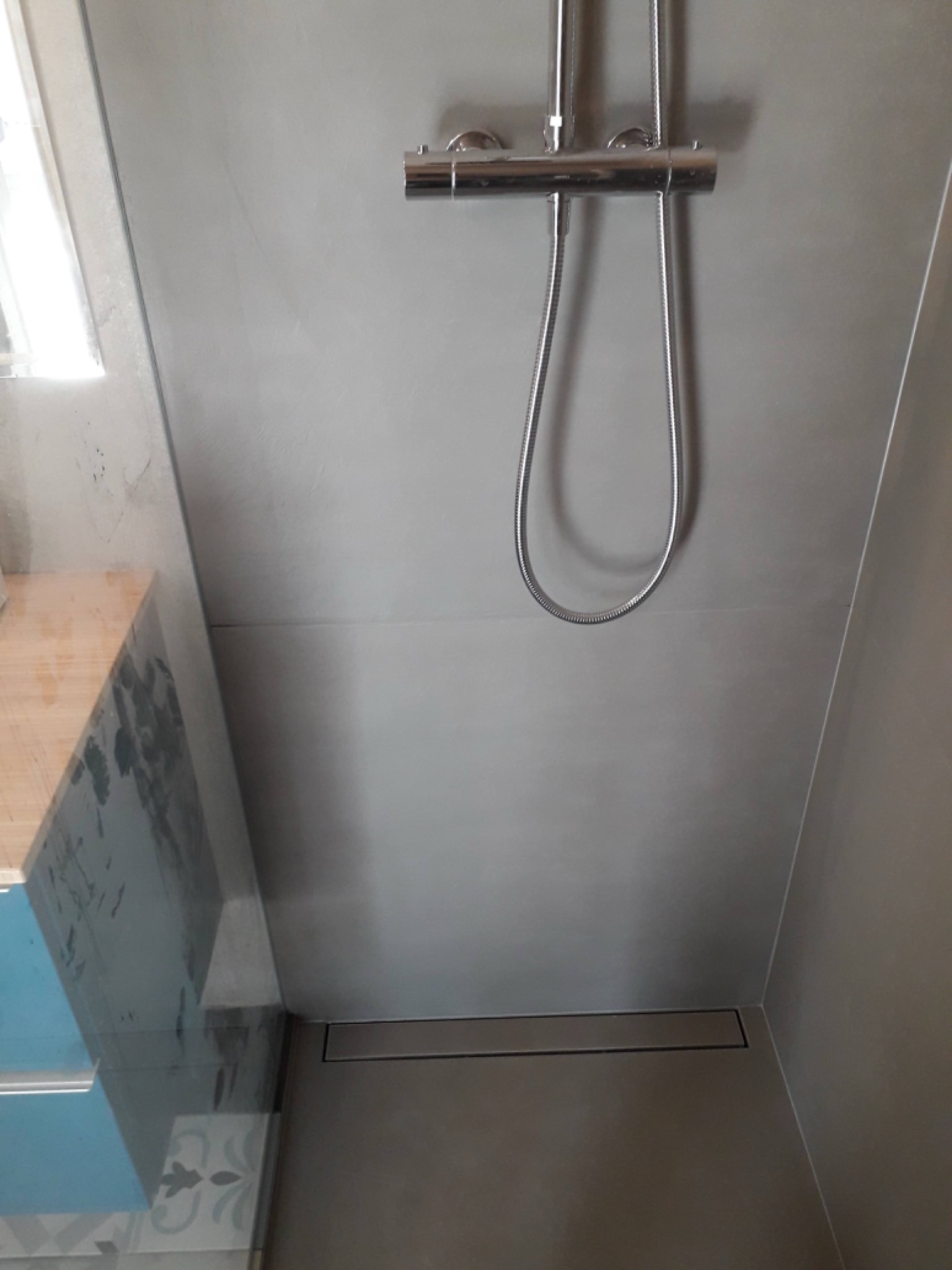 Szara kabina prysznicowa z odpływem liniowym w podłodze, widoczna bateria prysznicowa i fragment szafki z umywalką