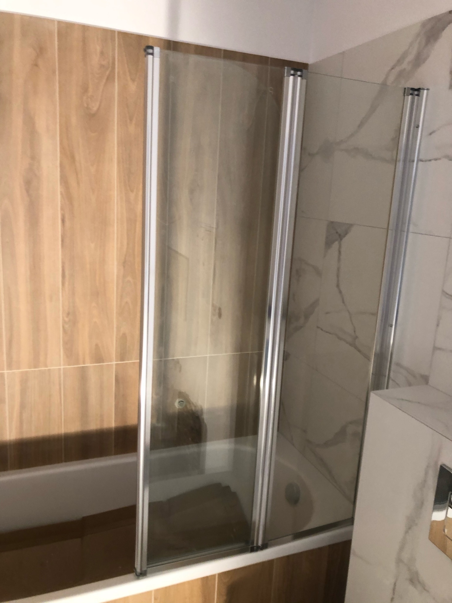 Szklany parawan nawannowy z chromowanymi profilami, zamontowany na wannie obłożonej drewnopodobnymi płytkami, widoczna ściana z płytek imitujących marmur.