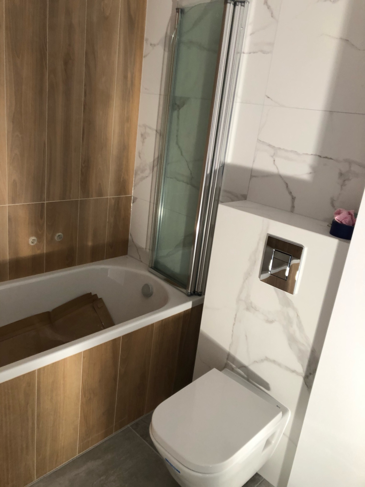 Nowoczesna łazienka z wanną obudowaną drewnopodobnymi płytkami, szklanym parawanem nawannowym i podwieszaną toaletą z marmurowym wykończeniem ściany.