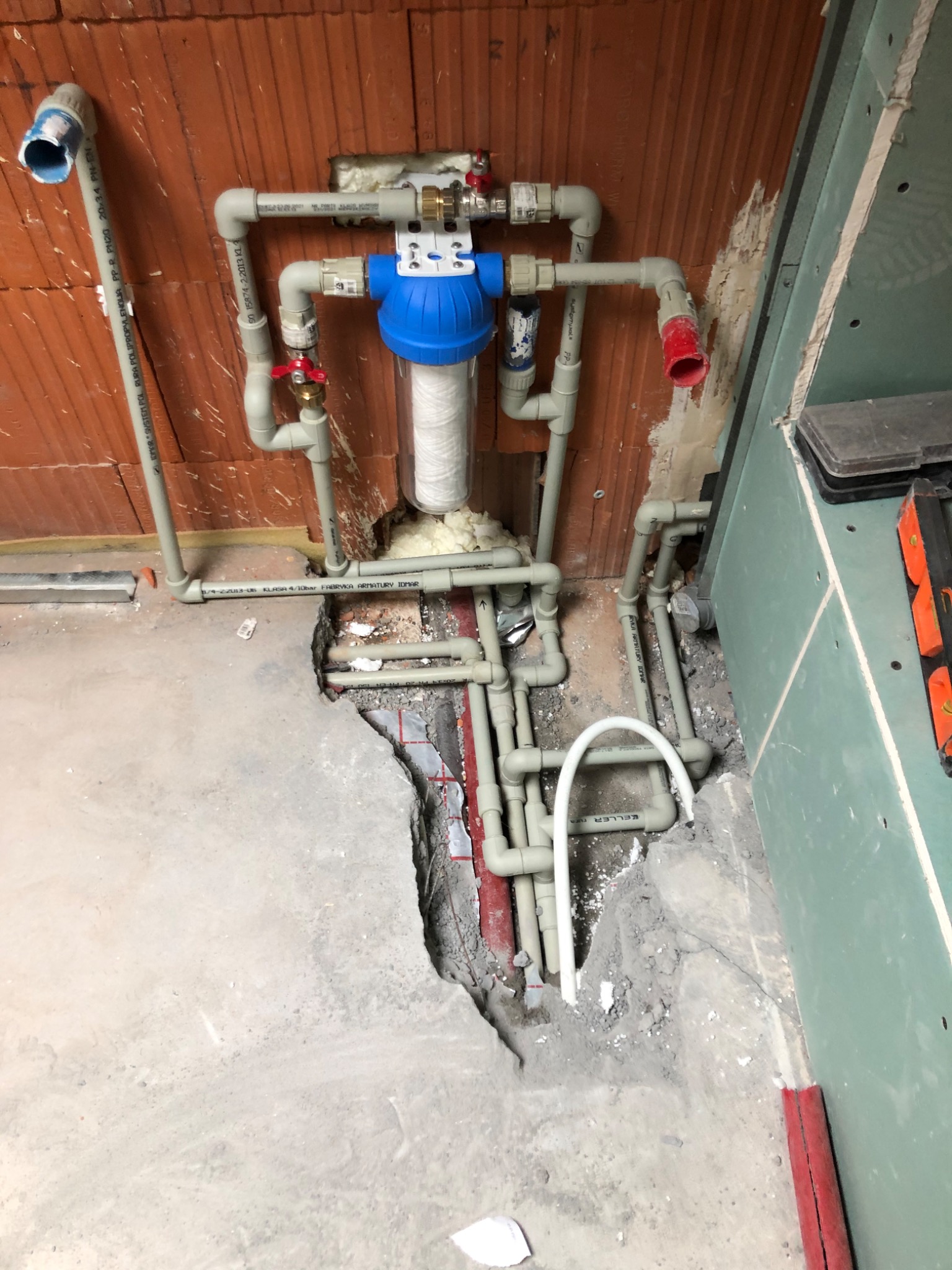 Montaż instalacji wodnej z filtrem w surowym stanie, widoczne rury z tworzywa sztucznego, ceglana ściana i płyta gipsowo-kartonowa.