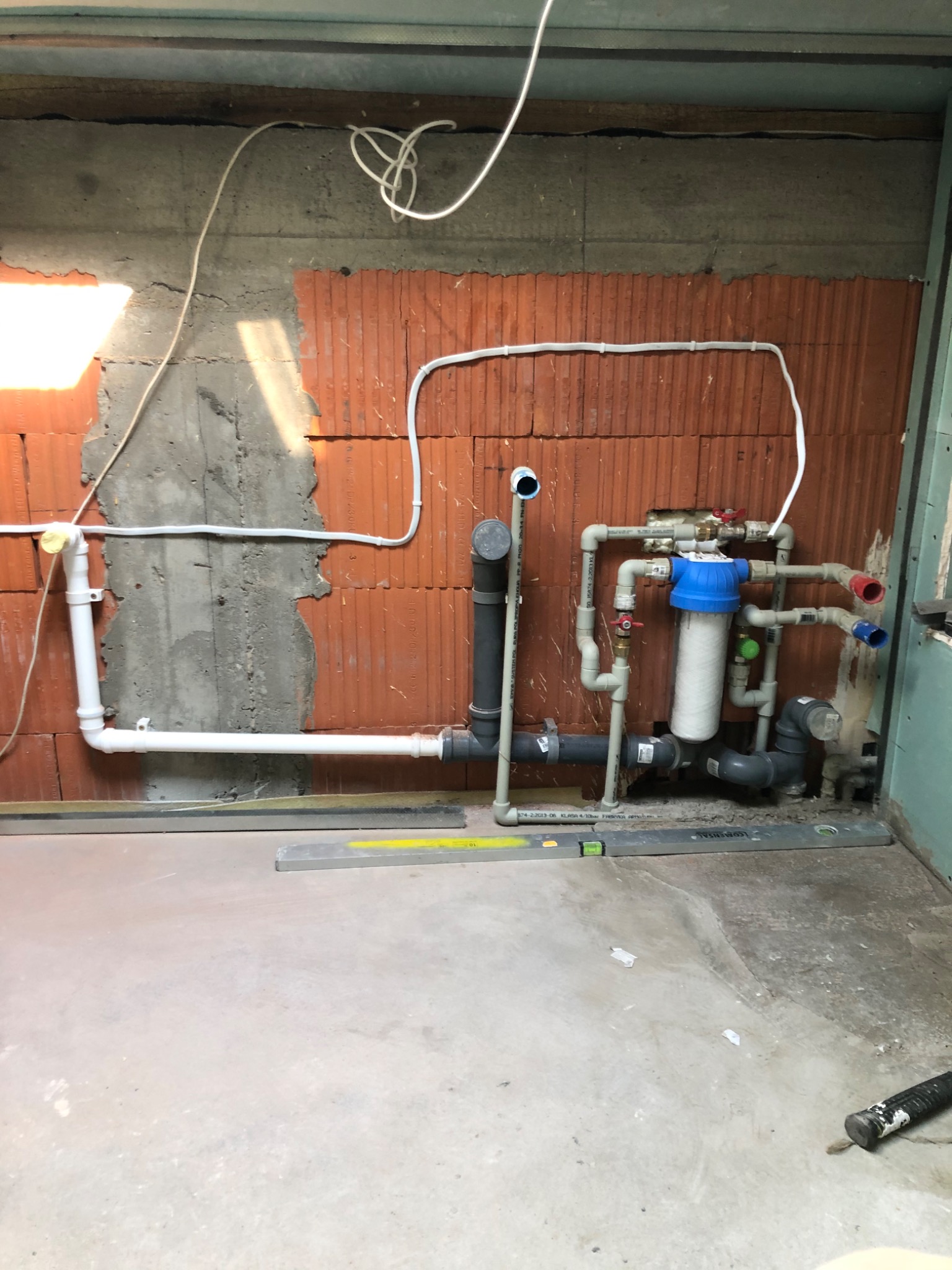 Surowa ściana z cegły i betonu z instalacją wodno-kanalizacyjną w trakcie montażu, widoczne rury PCV, filtry i poziomice na podłodze.