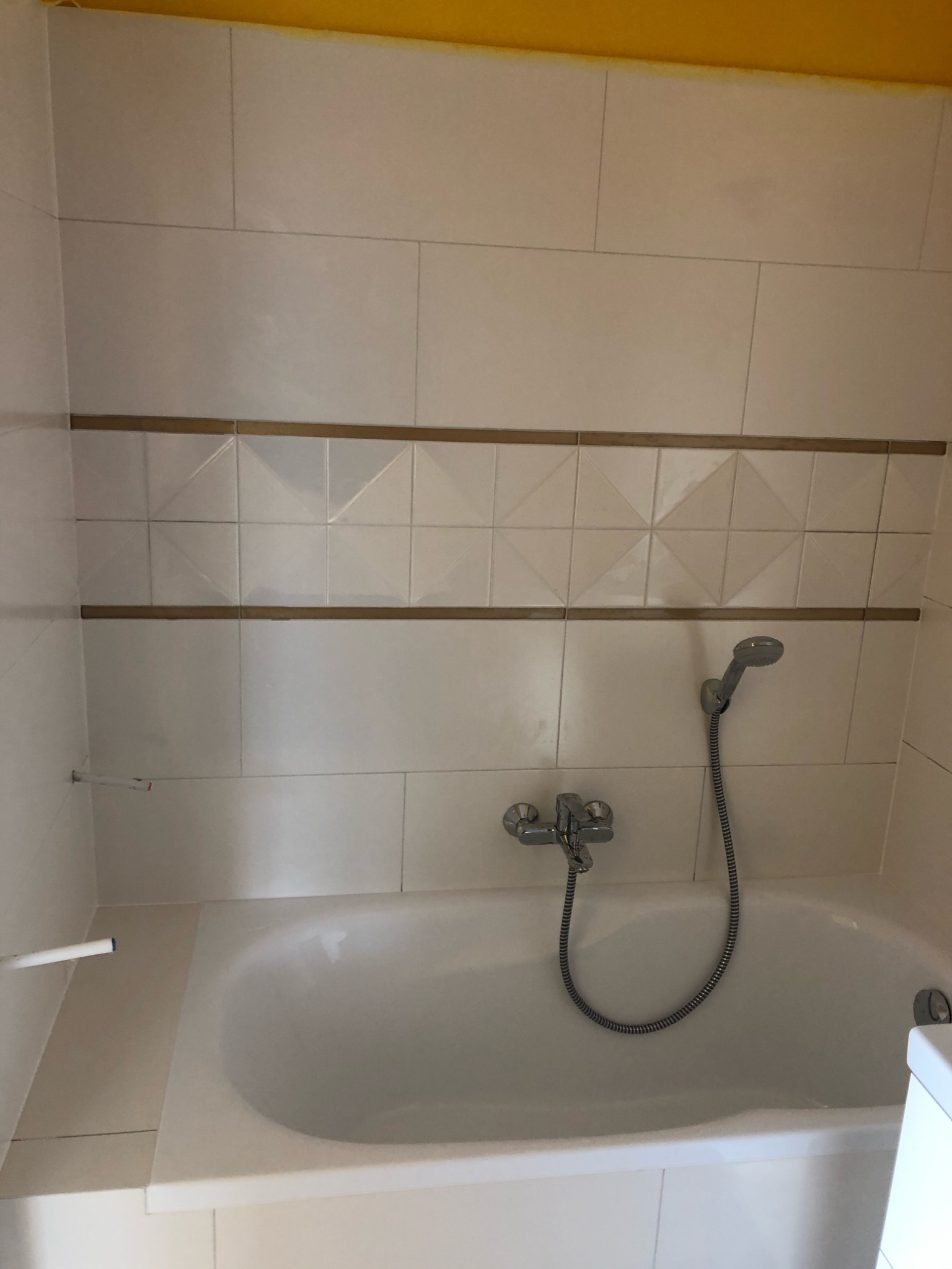 Biała wanna z chromowaną baterią i słuchawką prysznicową na tle ściany wyłożonej białymi płytkami z dekoracyjnym pasem z płytek z geometrycznym wzorem i żółtym sufitem.