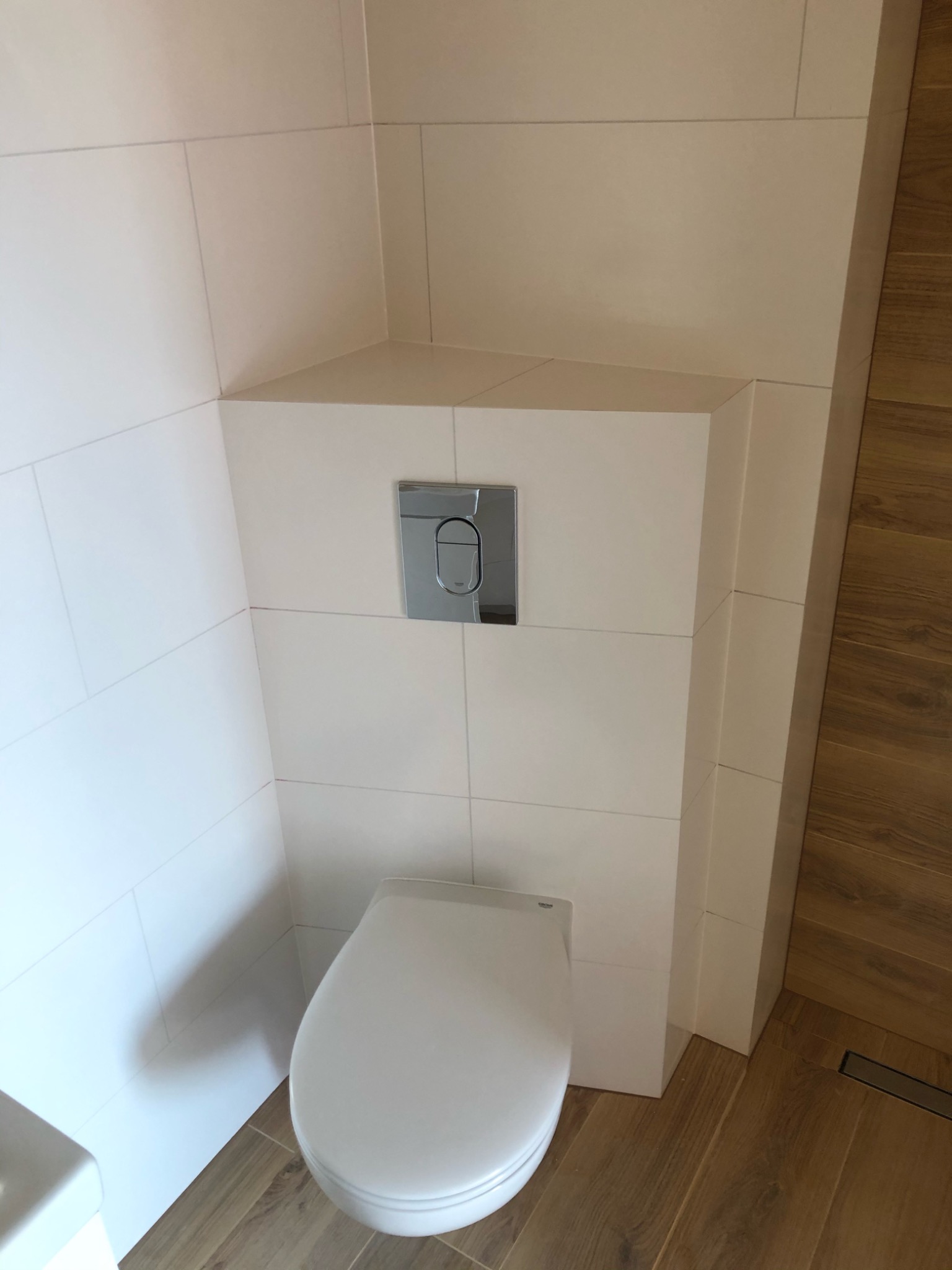 Nowoczesna toaleta podwieszana z chromowanym przyciskiem spłukującym, obudowana białymi płytkami ceramicznymi, z drewnopodobną podłogą i odpływem liniowym.