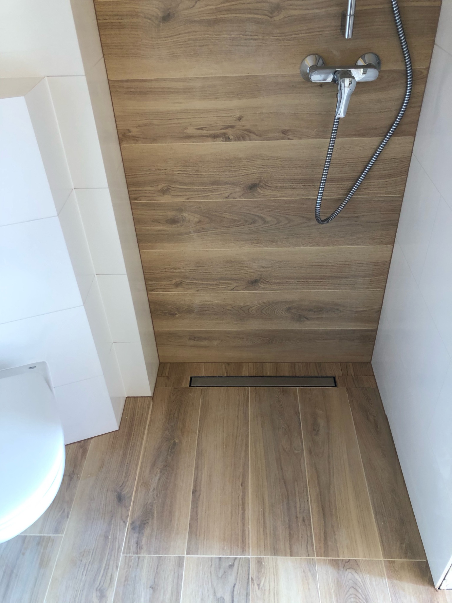 Nowoczesna kabina prysznicowa z drewnopodobnymi płytkami na ścianach i podłodze, odpływ liniowy, chromowana bateria natryskowa i fragment toalety z lewej strony.