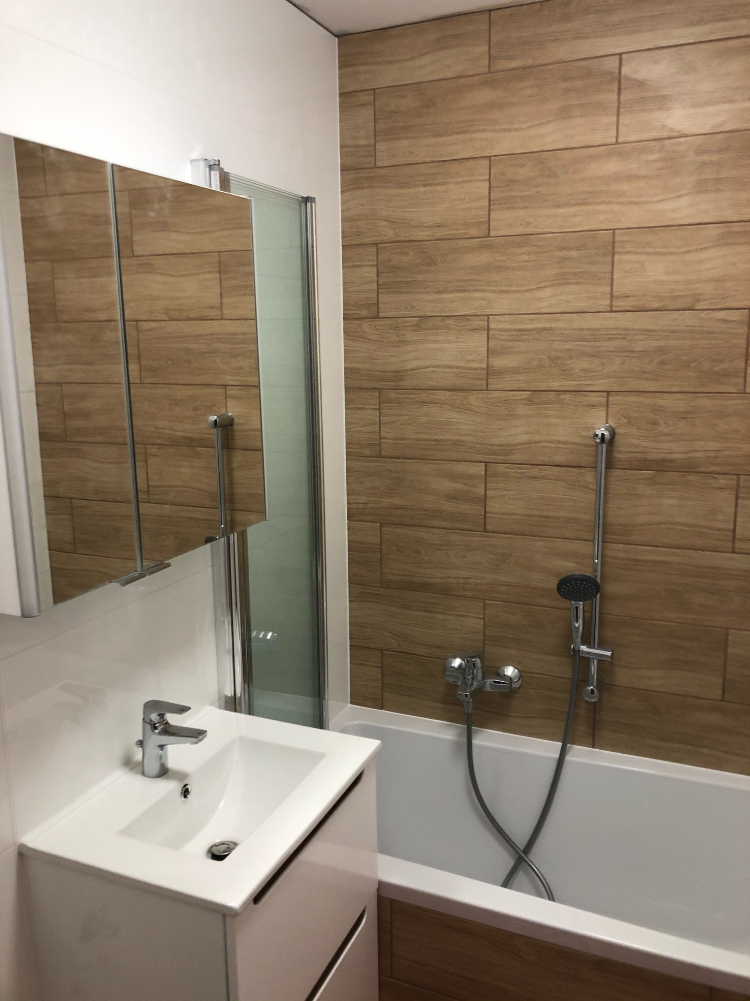 Nowoczesna łazienka z drewnopodobnymi płytkami, białą armaturą i lustrzaną szafką, widok na wannę ze słuchawką prysznicową.