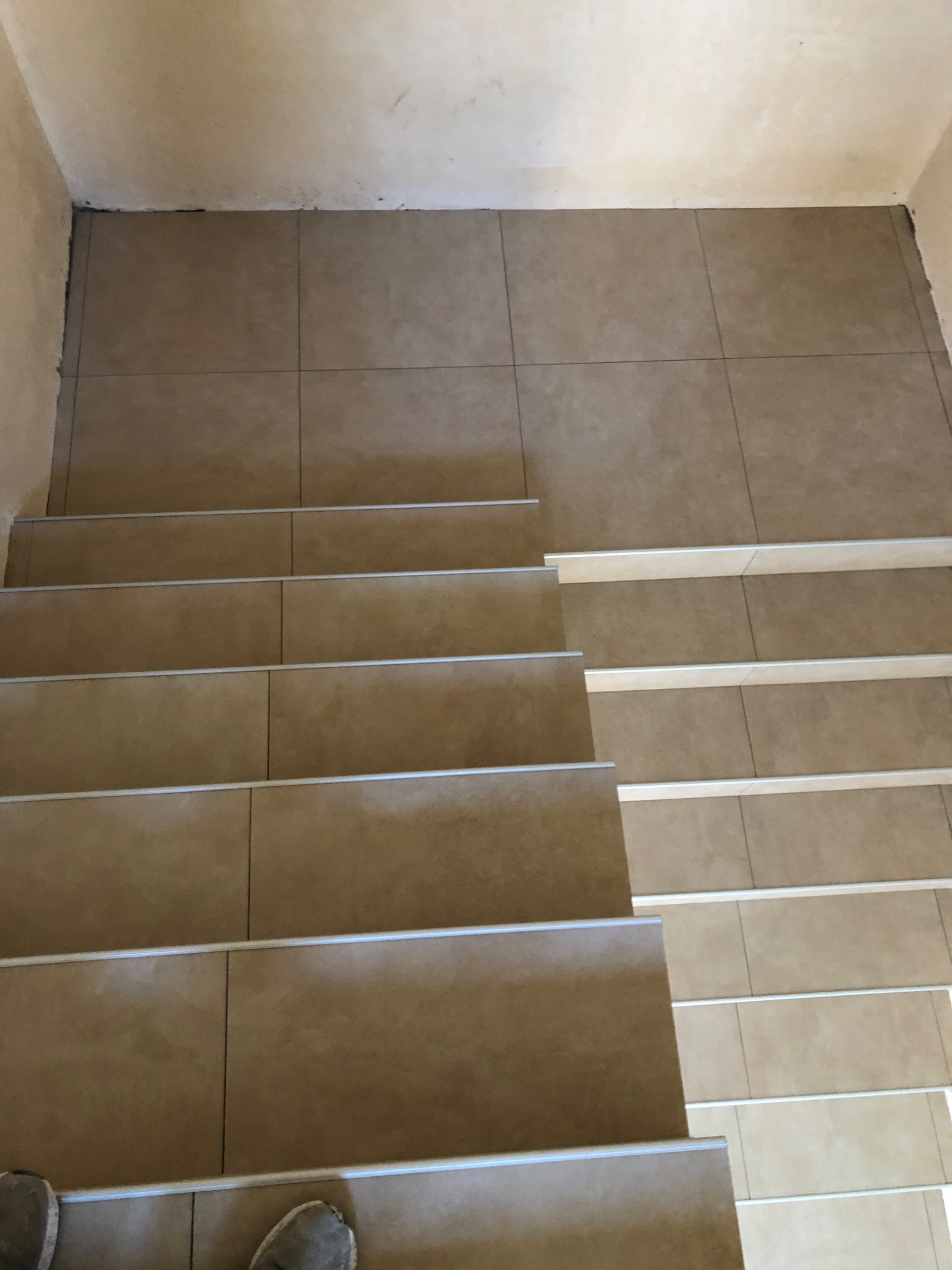 Wyłożone beżowymi płytkami schody z aluminiowymi profilami krawędziowymi, widoczne stopy w obuwiu w dolnej części kadru.