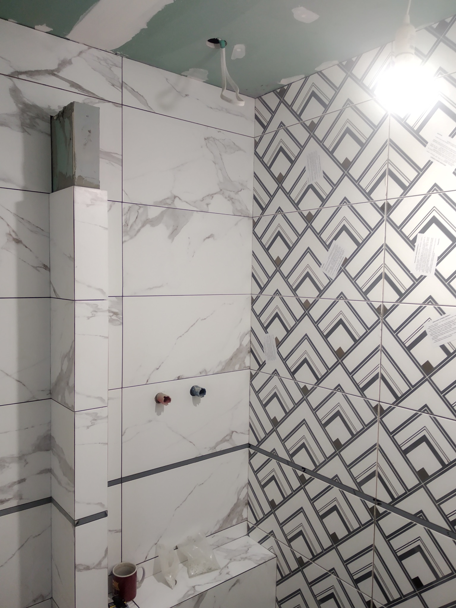 Ściana łazienki wyłożona białymi płytkami imitującymi marmur z szarymi żyłami, połączona z płytkami w geometryczne wzory, widoczne wystające rury i niedokończone wykończenie sufitu.