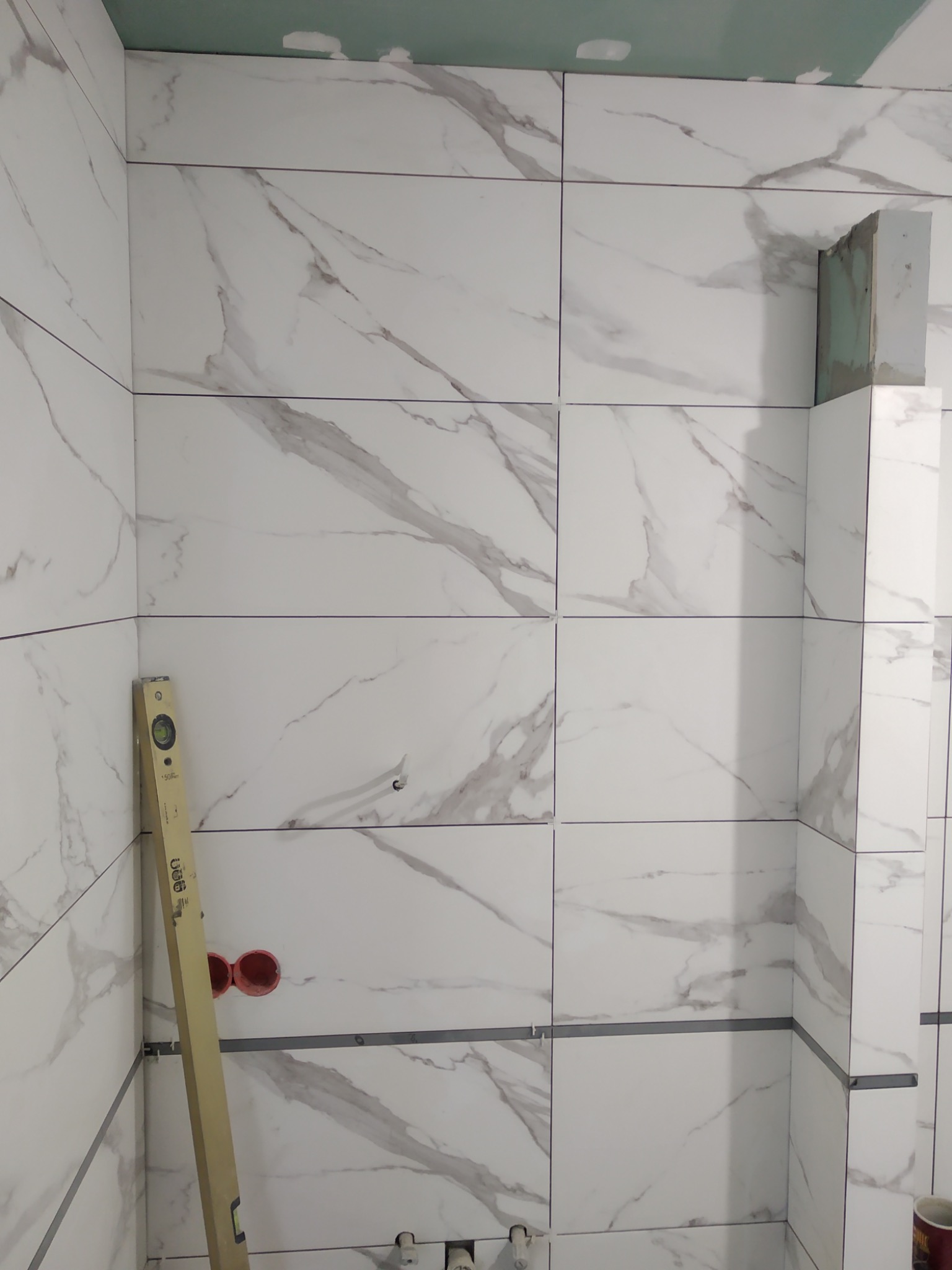 Ściana w trakcie układania płytek imitujących marmur, widoczny poziomica, otwory na instalacje i surowy fragment ściany w narożniku.