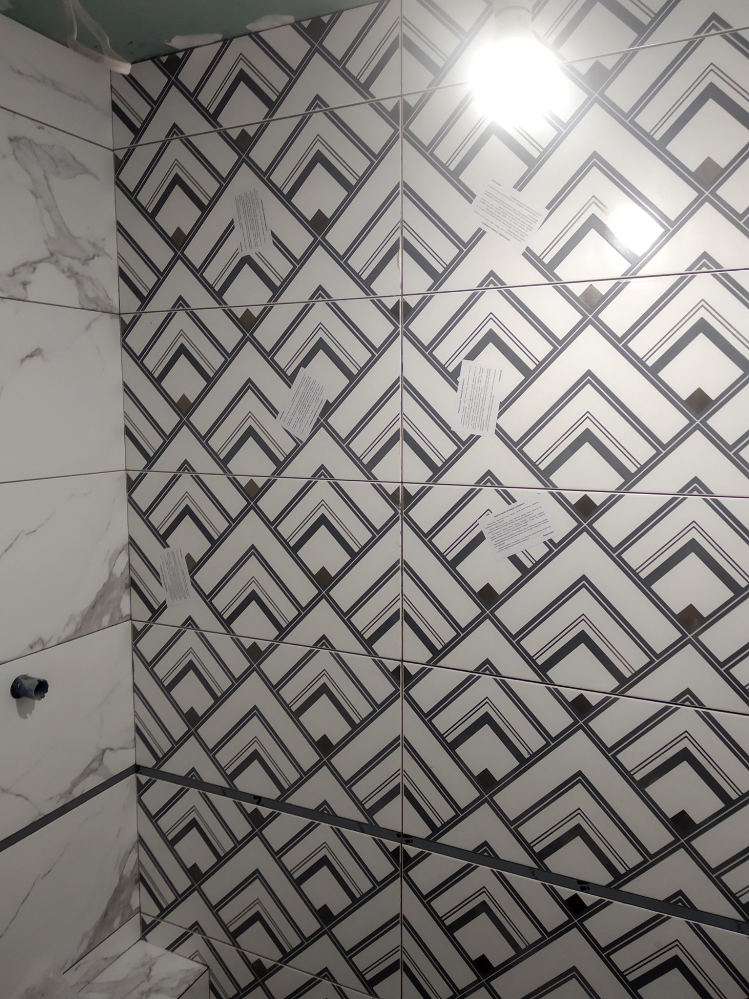 Ściana łazienki wyłożona białymi płytkami z szarym wzorem w strzałki, zabezpieczona taśmą malarską, z widocznymi fragmentami rur i przyklejonymi kartkami informacyjnymi.